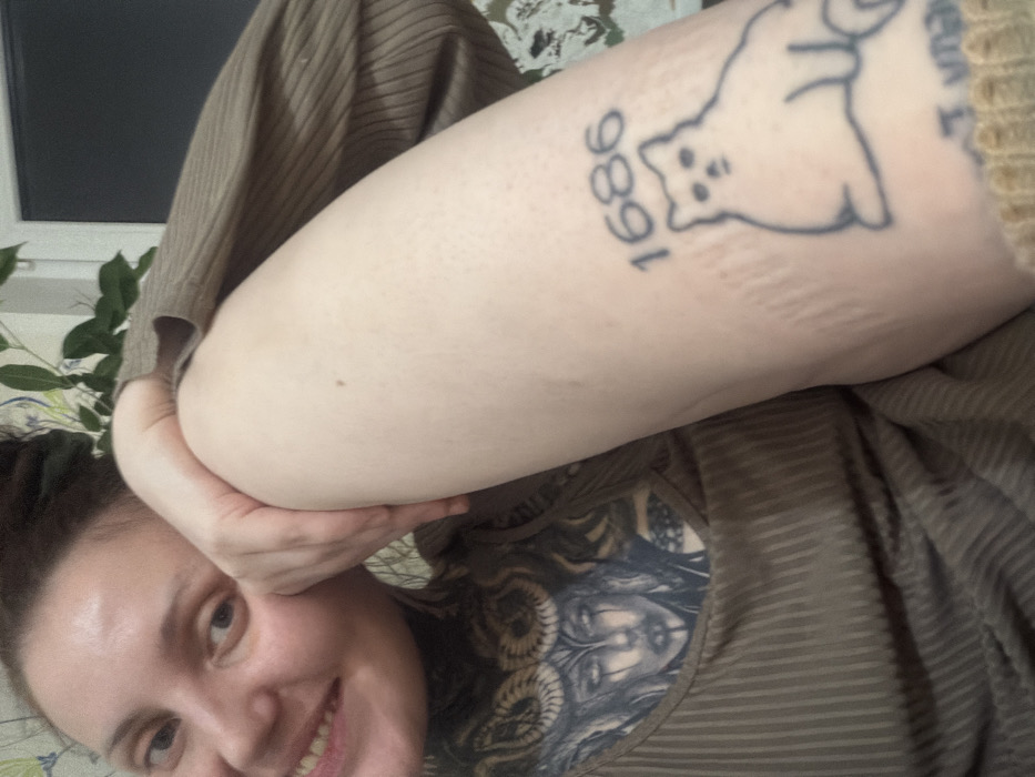 A smiling woman in a brown striped shirt holds her tattooed arm in the air. Her arm features a tattoo of the year "1686" beneath an outline of a cat. She has a visible chest tattoo with a pale face surrounded by snakes. A window, green plants, and floral wallpaper are in the background.