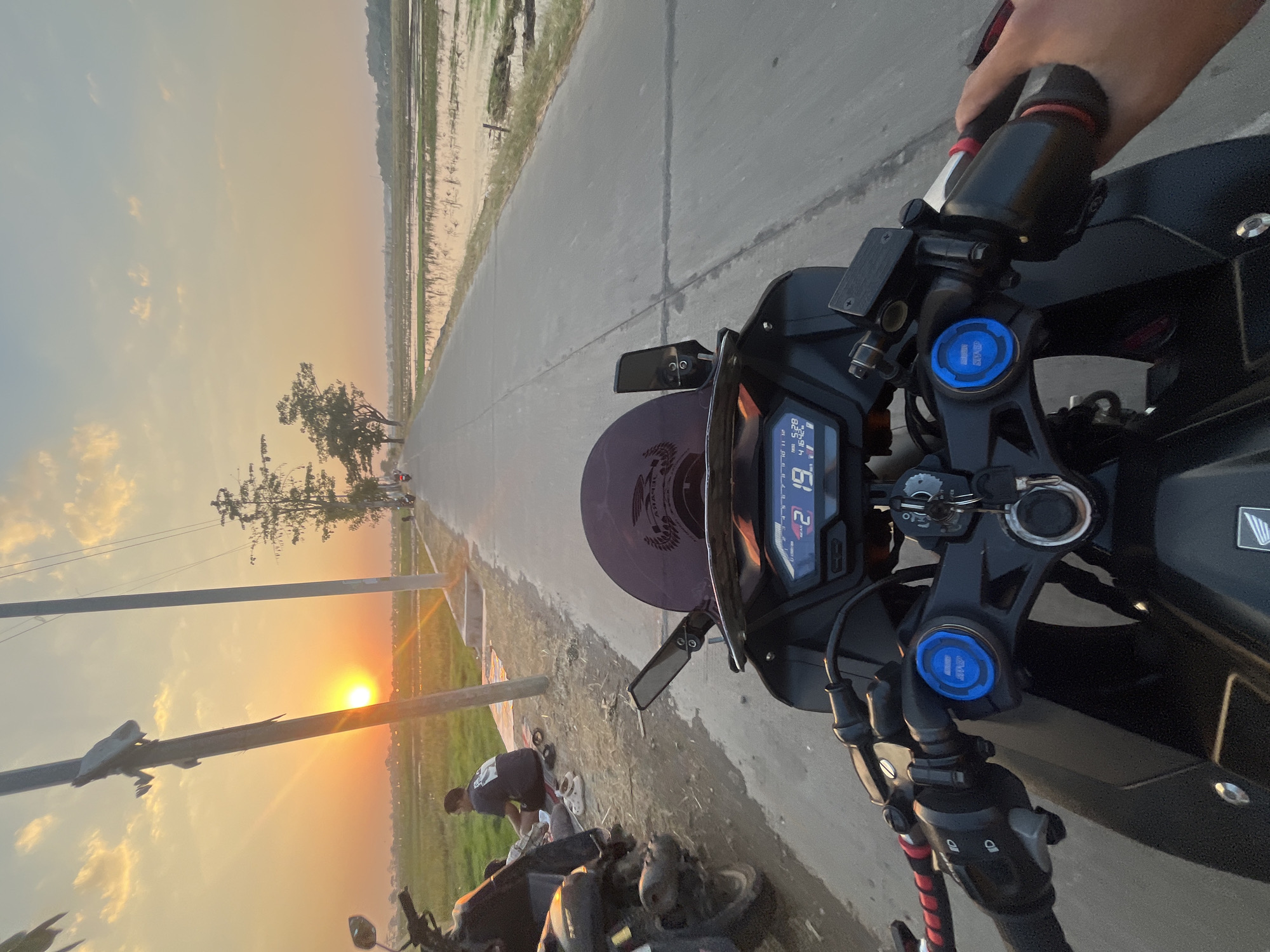 The image is a first-person perspective of someone riding a motorcycle on a paved road in Pecangaan, Indonesia, during sunset. The road runs through a rural landscape with rice paddies on the right and grassy fields on the left. The sun is low on the horizon, casting a warm, orange glow across the sky, illuminating scattered clouds.
In the foreground, the handlebars and dashboard of the motorcycle are visible, showing a digital display with readings that include speed (21 km/h) and time (5:28). A hand is gripping the right handlebar. Two other motorcycles are parked on the left side of the road, near where two people are sitting on the ground, possibly taking a break. The ground beside the road is covered with grass and some scattered debris. There are utility poles with wires running along the left side of the road. In the distance, more trees and hints of buildings can be seen, suggesting a scattered rural settlement. The overall scene depicts a peaceful moment of travel through a picturesque Indonesian countryside at dusk.