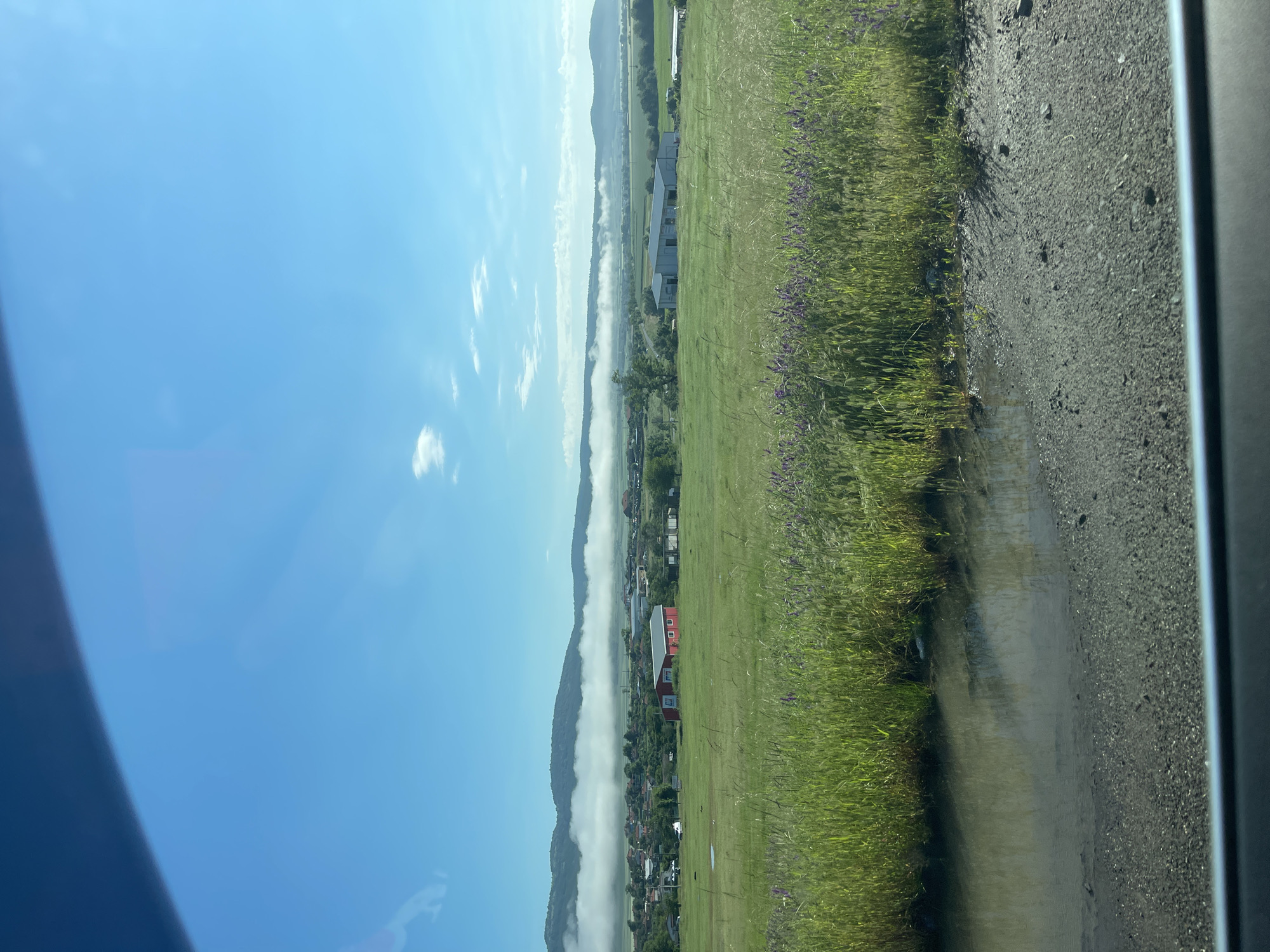 This image captures a panoramic view of a serene rural landscape, likely taken from a moving vehicle. The scene depicts a wide valley in Ruen, Bulgaria, under a clear, bright blue sky dotted with scattered white clouds.
In the foreground, a strip of vibrant green grass mixed with purple wildflowers runs along the bottom left. Adjacent to it on the right is a rough dirt or gravel road, partially wet or reflective in areas, suggesting recent rain or morning dew. A dark vertical bar on the far right edge indicates the frame of a window, possibly from a car.
The mid-ground is dominated by a striking band of low-lying fog or mist, stretching horizontally across the valley floor. This fog partially obscures houses and other buildings nestled within the valley, creating a layered effect. Above the fog bank, on the left, distant green mountains or hills rise, their peaks reaching towards the sky. Below the fog, more signs of human habitation, including a distinctive red-roofed building and a modern grey structure, are visible among trees and fields.
The presence of low-lying fog under a bright sky strongly suggests an early morning scene, shortly after sunrise, as the mist begins to dissipate. No people are visible, and there is no visible text. The overall impression is one of tranquil natural beauty in a rural setting.