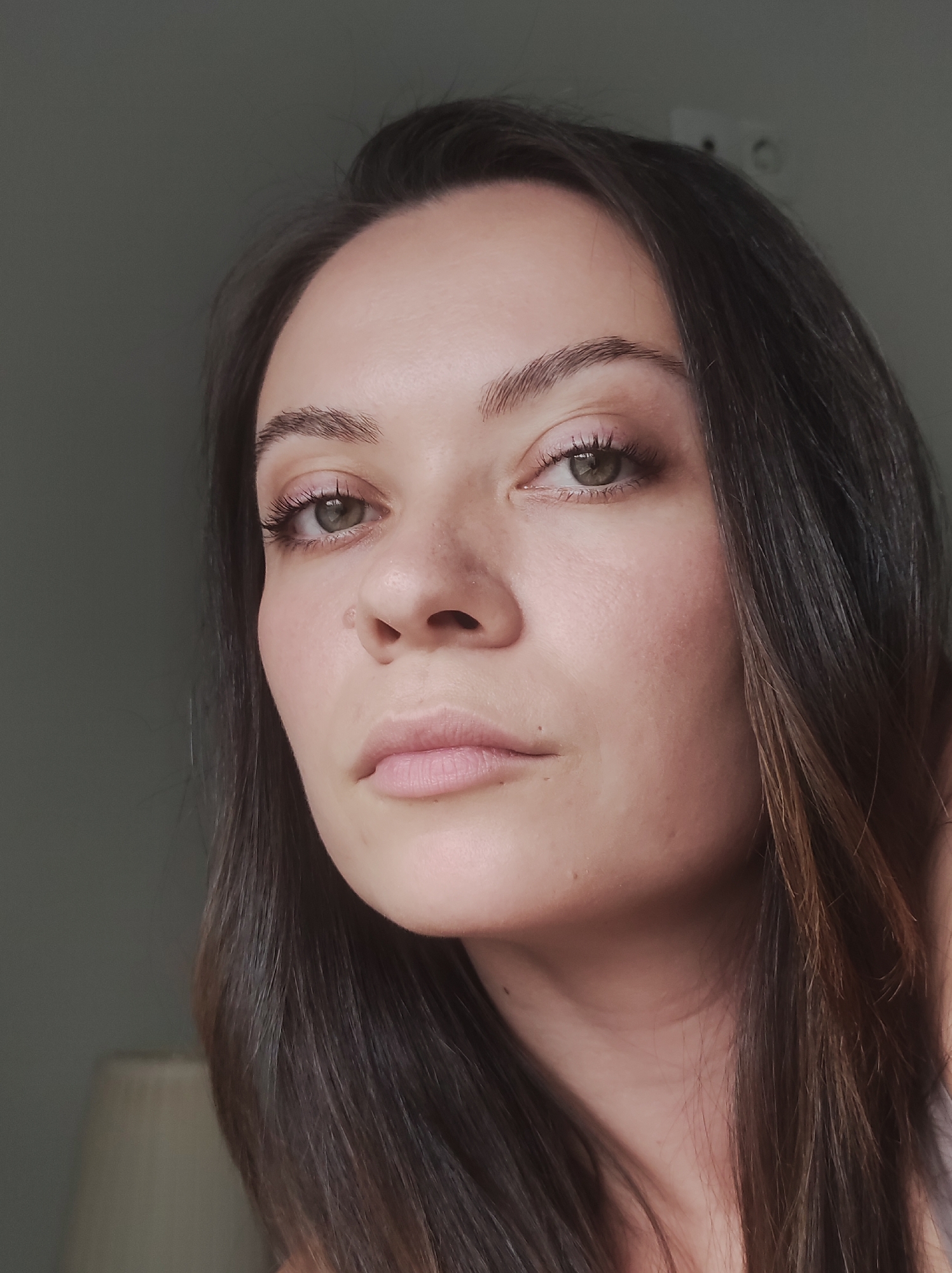 A close-up portrait shows a woman with light skin, brown hair with subtle highlights, and green eyes. She faces the camera directly with a serious expression. Her lips are a light pink color, and her eyebrows are groomed and natural. The background is a blurred, indistinct gray, with a hint of a light-colored lamp in the lower left corner. There are no identifiable markers to definitively place the image in Chaikovsky, Russia. The scene appears to be indoors.