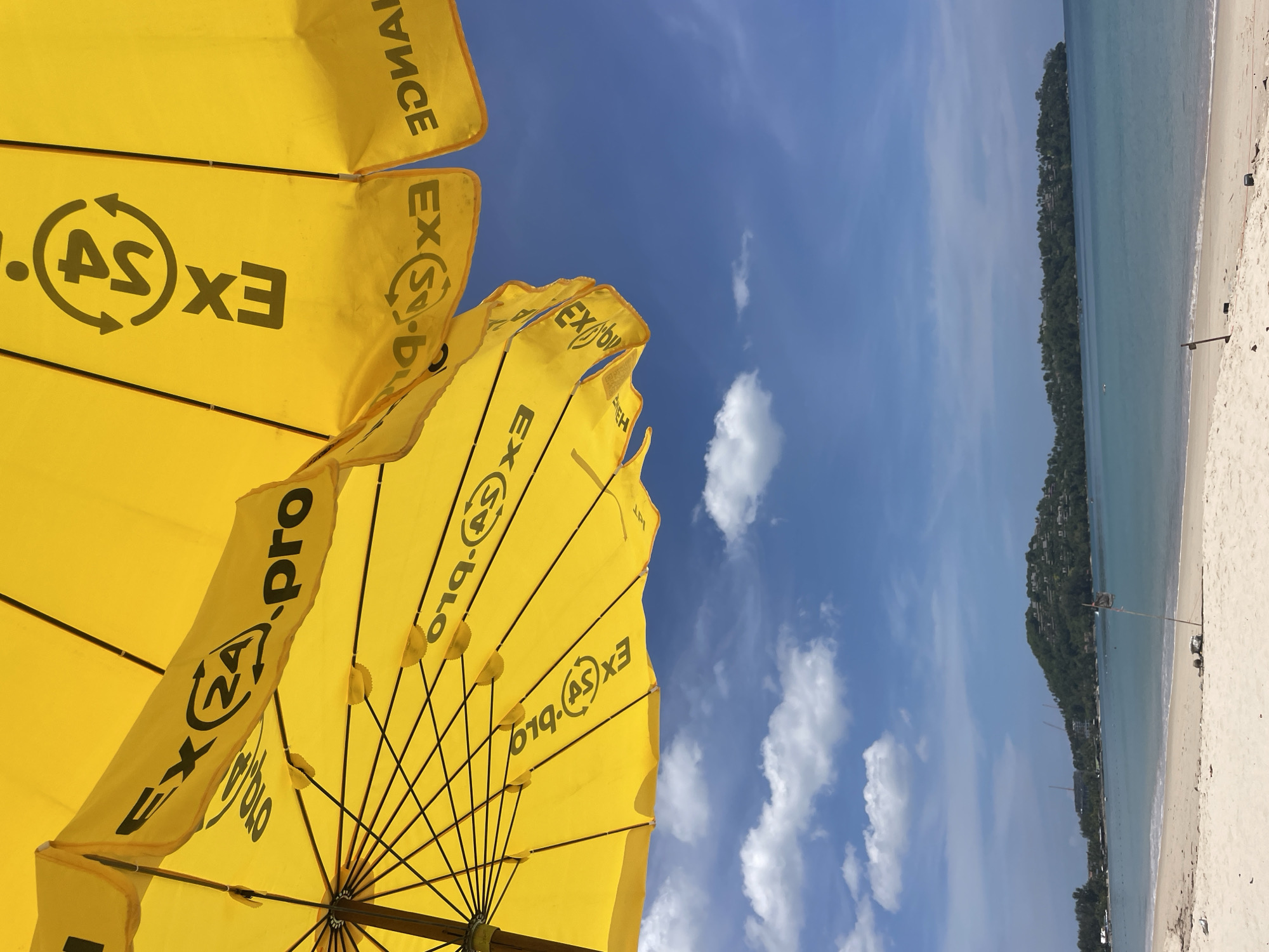 This image captures a vibrant and serene beach scene, looking out from beneath a cluster of yellow beach umbrellas. The setting is likely Choeng Thale, Thailand, suggested by the tropical landscape.
In the foreground, several bright yellow umbrellas dominate the view, providing a stark contrast against the blue sky. The underside of the umbrellas reveals their black radial spokes. Printed repeatedly on the umbrella fabric, in black capital letters, is text that appears to be "EX.S.pro" or a variation thereof, sometimes mirrored or partially obscured by folds. A distinctive logo featuring a circular arrow design encircles what looks like an "S" or "AS" within the text.
Beyond the umbrellas, a vast expanse of clear, azure sky is visible, dotted with fluffy white clouds, indicating sunny weather. The bright illumination suggests it is midday or early afternoon.
To the right, a pristine, light-colored sandy beach stretches alongside calm, turquoise-blue ocean waters. The waves appear gentle, lapping softly at the shore. In the distance, a lush, green hillside covered in dense tropical vegetation forms the horizon, with what appear to be buildings or resorts nestled amongst the trees. The overall scene evokes a sense of peace and a perfect tropical vacation atmosphere. No specific human activity is clearly depicted, but the presence of beach umbrellas implies people are nearby, enjoying the sun and shade.