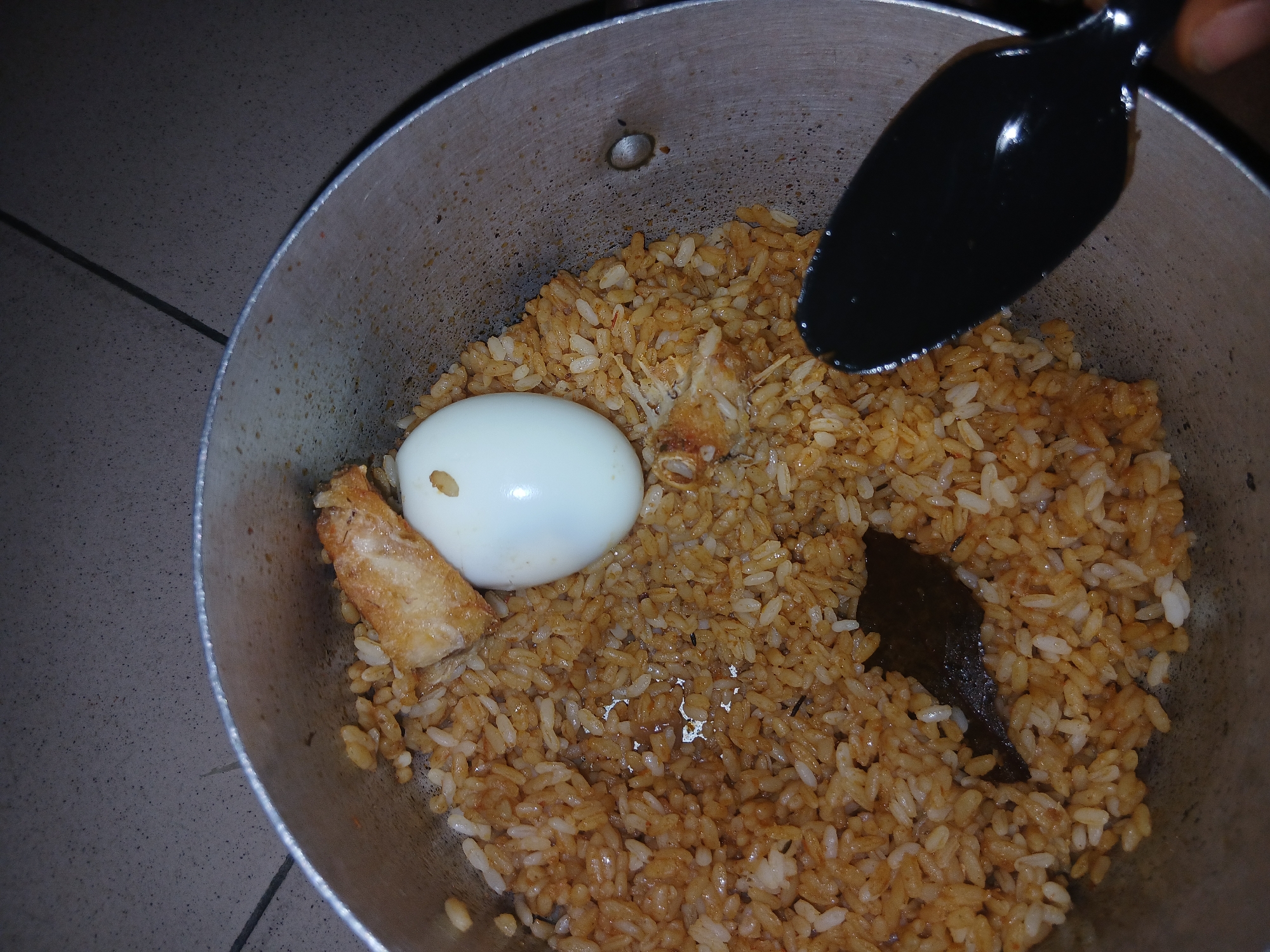 A close-up, overhead shot shows a pot of cooked rice with some additions. The rice appears to be a light brown color, likely from being seasoned or cooked in a broth. Two pieces of fried chicken or meat are visible, one larger piece with crispy skin, and a smaller, shredded piece near a hard-boiled egg. The egg is peeled and sits whole in the pot. A black spoon is positioned over the rice, with its handle extending out of the frame and its bowl hovering above the food, suggesting someone is about to serve or stir it. The pot itself is made of metal and appears somewhat worn. The background is a tiled surface, likely a floor, in muted tones of gray and brown. The lighting is dim, possibly indicating it's nighttime or an indoor setting with low light. The scene focuses solely on the food within the pot and the serving utensil. There is no visible text in the image.