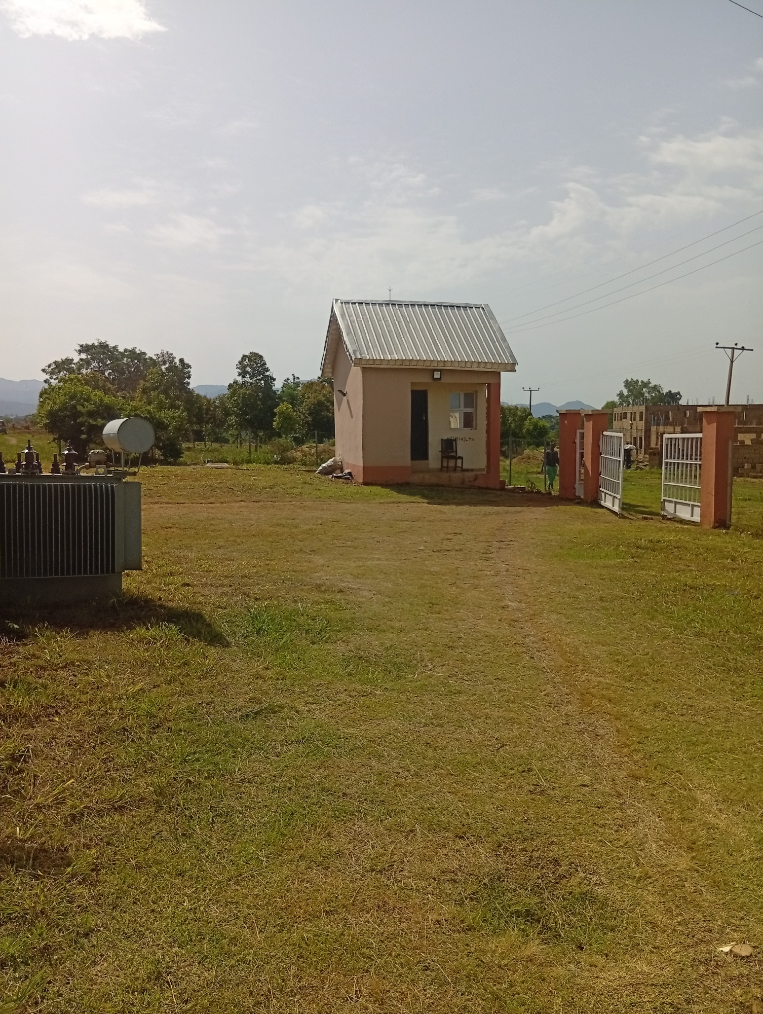 This is a daytime, eye-level, outdoor, full shot image taken in the city of Gwong, Nigeria. The scene depicts a small, tan-colored building with a corrugated metal roof, resembling a security post or small office, situated in a grassy field. A large, grey electrical transformer is visible to the left of the building. In the background, there is a line of trees, and further in the distance, there are unfinished brick buildings under construction and a power pole with lines. To the right of the main building, a set of open, white metal gates with brick pillars suggests an entrance to the property. One person, wearing a green shirt and light-colored pants, is walking near the gates. The sky is mostly clear with a few scattered clouds. The overall environment appears to be a rural or semi-developed area.