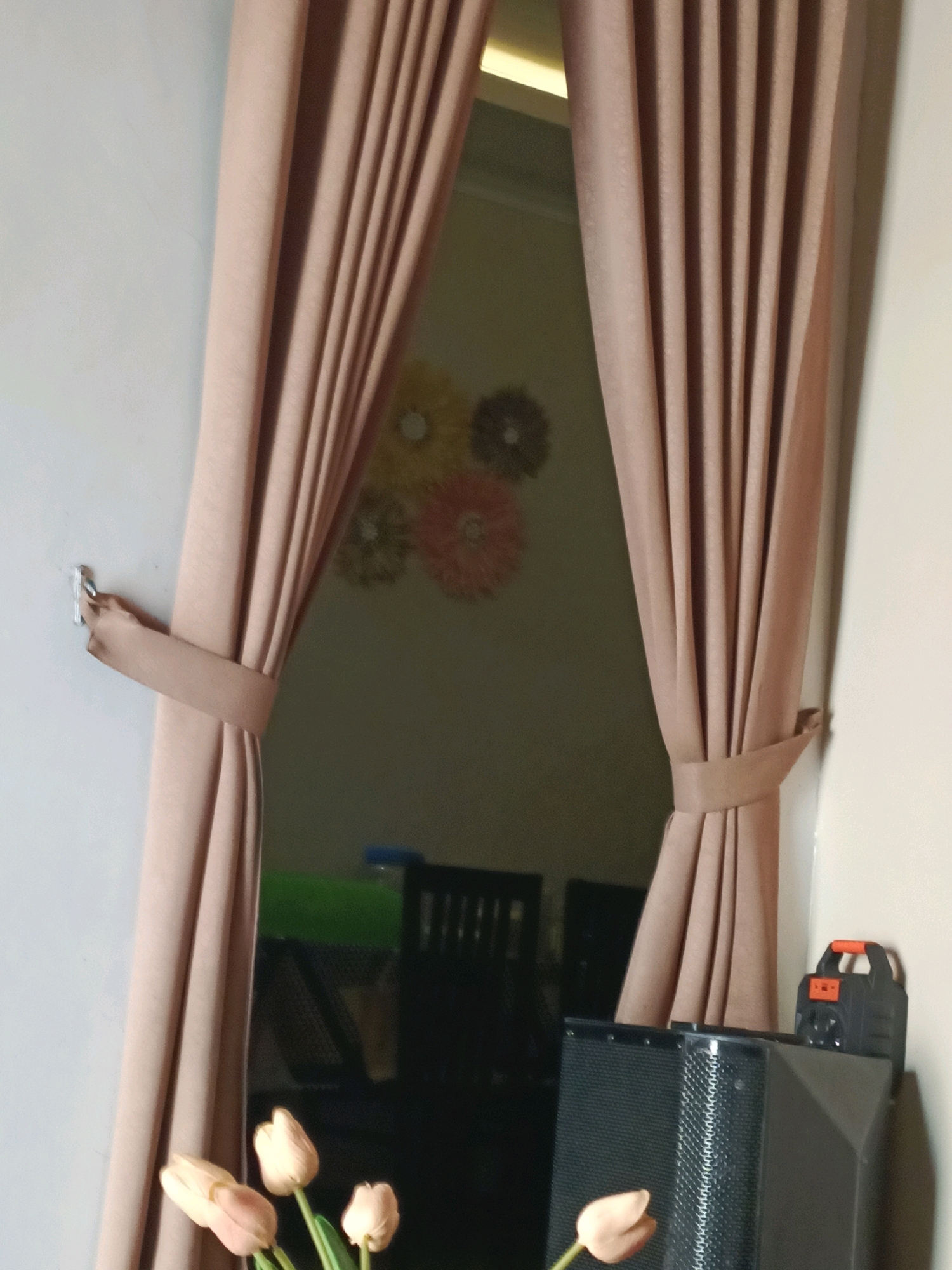 The image depicts an indoor scene with a window covered by draped, tan-colored curtains. The curtains are tied back on both sides with matching fabric bands. Through the gap in the curtains, a dimly lit room is visible, featuring a wall with floral decorations and furniture that is not clearly discernible. In the foreground, on the right side of the image, a black speaker is present, topped by a small black and orange portable power bank. In the lower left corner, several pale pink tulip buds are visible. The overall impression is that of a domestic interior, possibly a living room or a similar space. The mention of Rancang Kencono, Indonesia, suggests a specific geographical location for this scene, but the image itself does not contain any visual cues to confirm or deny this location.