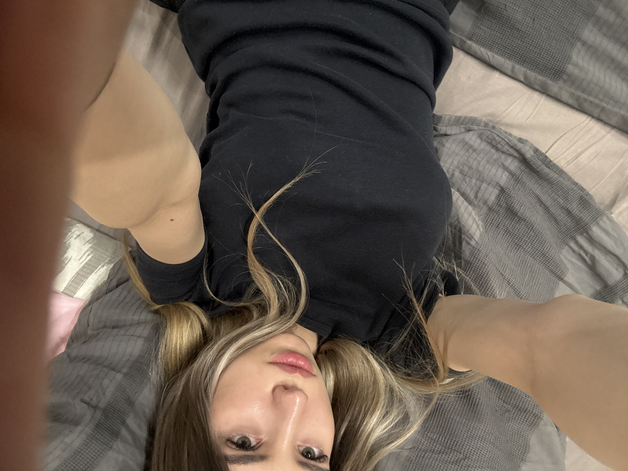 A person is lying on their back on a bed covered with a gray and beige patterned duvet. They are wearing a black long-sleeved shirt. The person's face is visible, showing blonde hair with dark roots, eyes, nose, and lips. The person's arms are raised on either side of their head, with one arm more prominent in the foreground. The background shows the bed linen and a portion of a pillow. There is no information in the image to support the location of Chaikovsky, Russia.