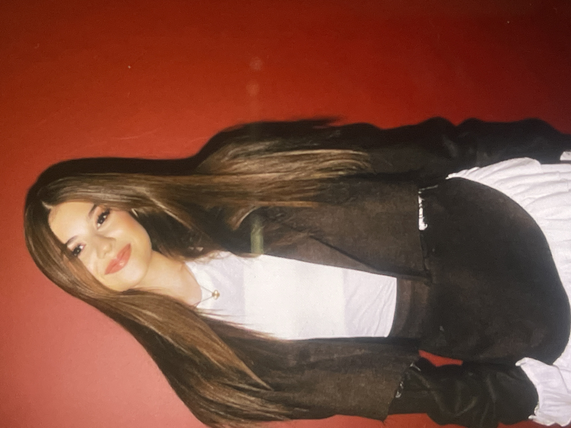 The image, rotated 90 degrees clockwise, captures a young woman posing for a photo against a plain red background. She is looking directly at the camera with a warm, open-mouthed smile, revealing a confident and happy expression. Her long, straight, dark brown hair, parted in the middle, cascades around her shoulders and spreads out behind her.
She is wearing a simple white t-shirt underneath a loose-fitting, dark (likely black or very dark brown) long-sleeved top or jacket. A delicate gold-colored chain with a small pendant is visible around her neck. Her makeup includes dark eyebrows, subtle eye makeup, and a vibrant red-orange lipstick that enhances her smile.
The background is a solid, deep red or dark orange surface, possibly a wall, with a few faint specks or imperfections visible upon closer inspection. The lighting appears soft and even, suggesting an indoor setting. The overall image has a slightly soft focus and a subtle graininess, contributing to a casual aesthetic. There is no visible text in the image. The photo provides no specific location cues beyond the plain background, but the context indicates it was taken in Poltava, Ukraine.