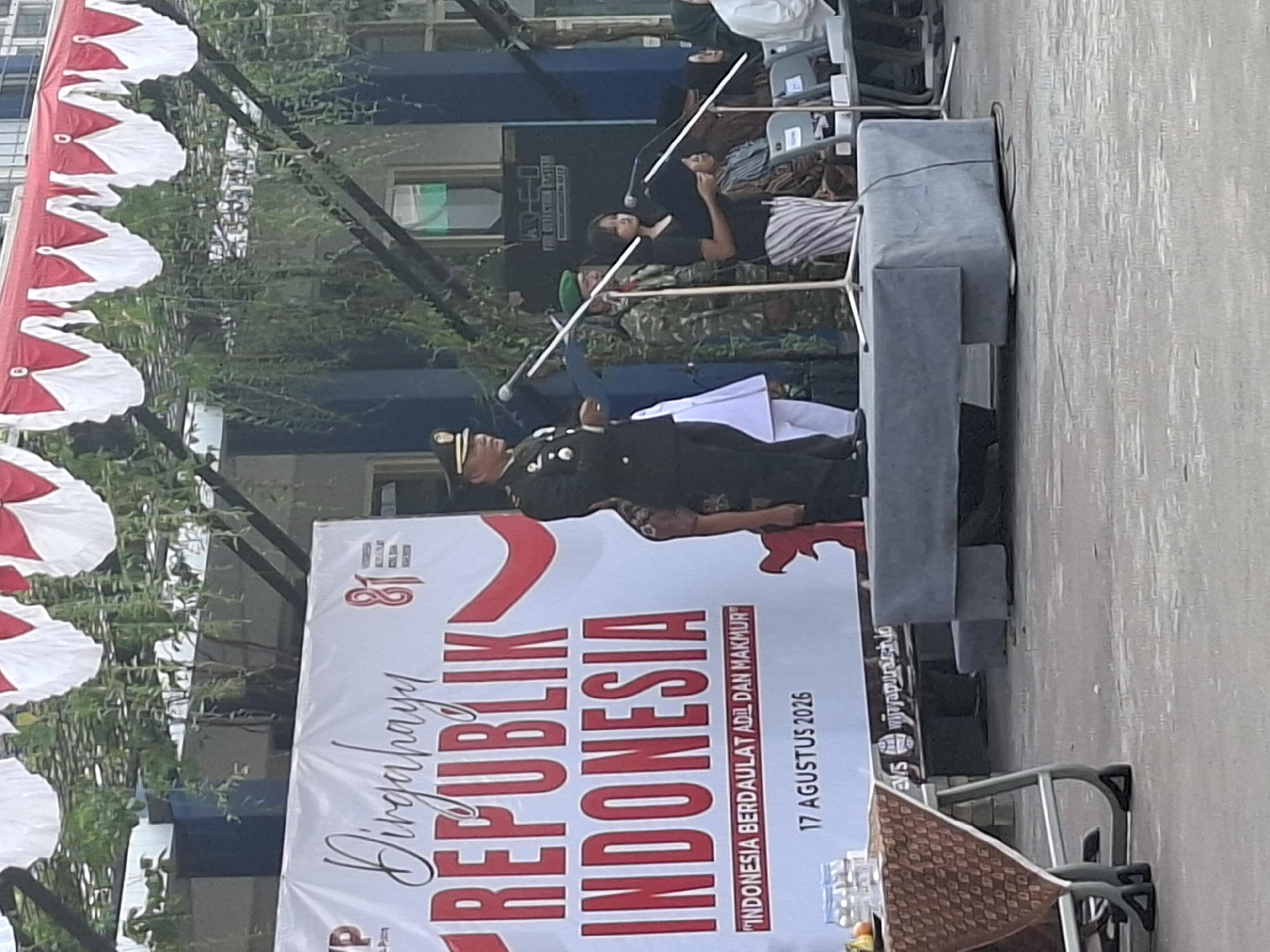 The image depicts an outdoor ceremony in Surabaya, Indonesia, likely at a government building or public space, given the patriotic banner and uniformed officials. A large white banner with red lettering prominently displays "DIRGAHAYU REPUBLIK INDONESIA" (Happy Indonesian Republic) and "INDONESIA BERDAULAT ADIL DAN MAKMUR" (Sovereign, Just, and Prosperous Indonesia), dated "17 AGUSTUS 2026." A man in a military uniform is speaking at a microphone on a raised platform, with several other people, including a woman holding a microphone and others in military attire and civilian clothing, present. The setting appears to be a semi-public gathering, with a table draped in a patterned cloth to the left, and seating areas visible in the background. The red and white bunting draped above suggests a national holiday celebration. The overall scene conveys a formal and patriotic atmosphere.