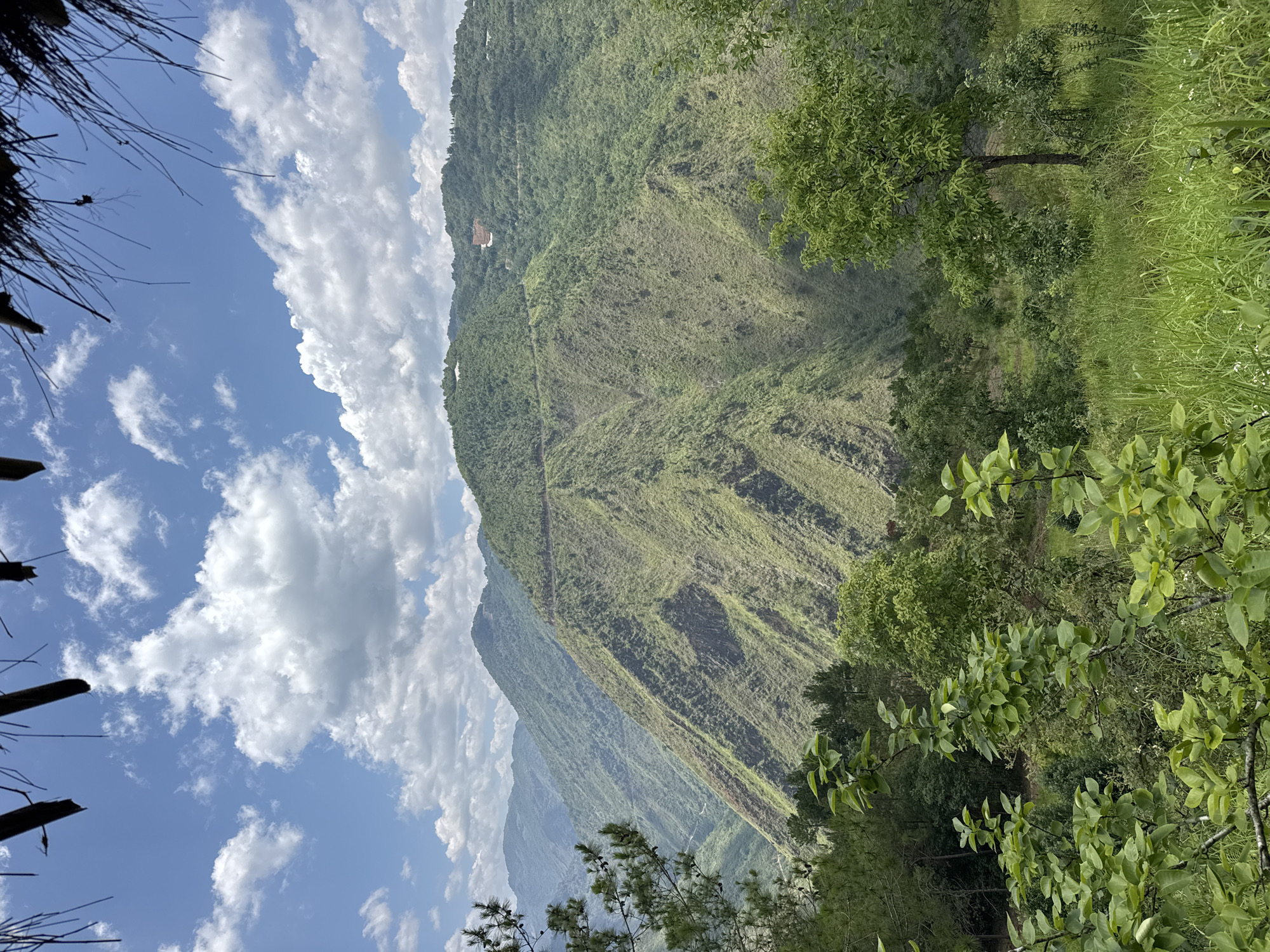 The image depicts a landscape view taken from a high vantage point in Lũng Cú Commune, Vietnam. The scene showcases a steep, green-covered mountain range with a few buildings near the summit. The sky is blue with puffy white clouds. In the foreground, there are patches of green vegetation and foliage. A thatched roof structure is visible in the upper left corner of the frame.