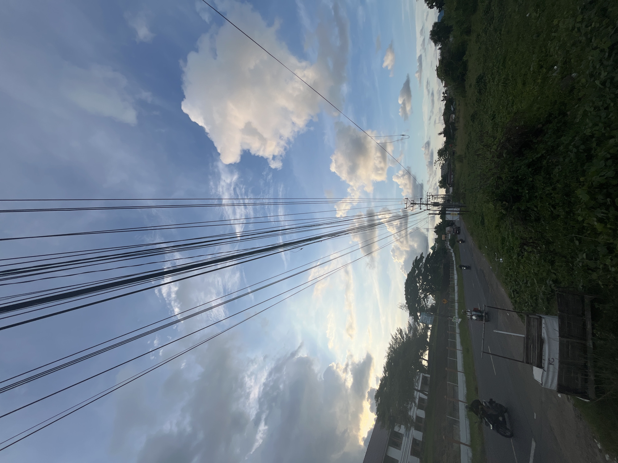The image depicts a street scene in Lhokseumawe, Indonesia. A road stretches into the distance, flanked by trees and dense vegetation. Several motorcycles are visible on the road. Overhead, a network of power lines crisscrosses the sky, which is filled with scattered clouds. A building is partially visible on the lower left side of the image. A cart or structure is on the side of the road.