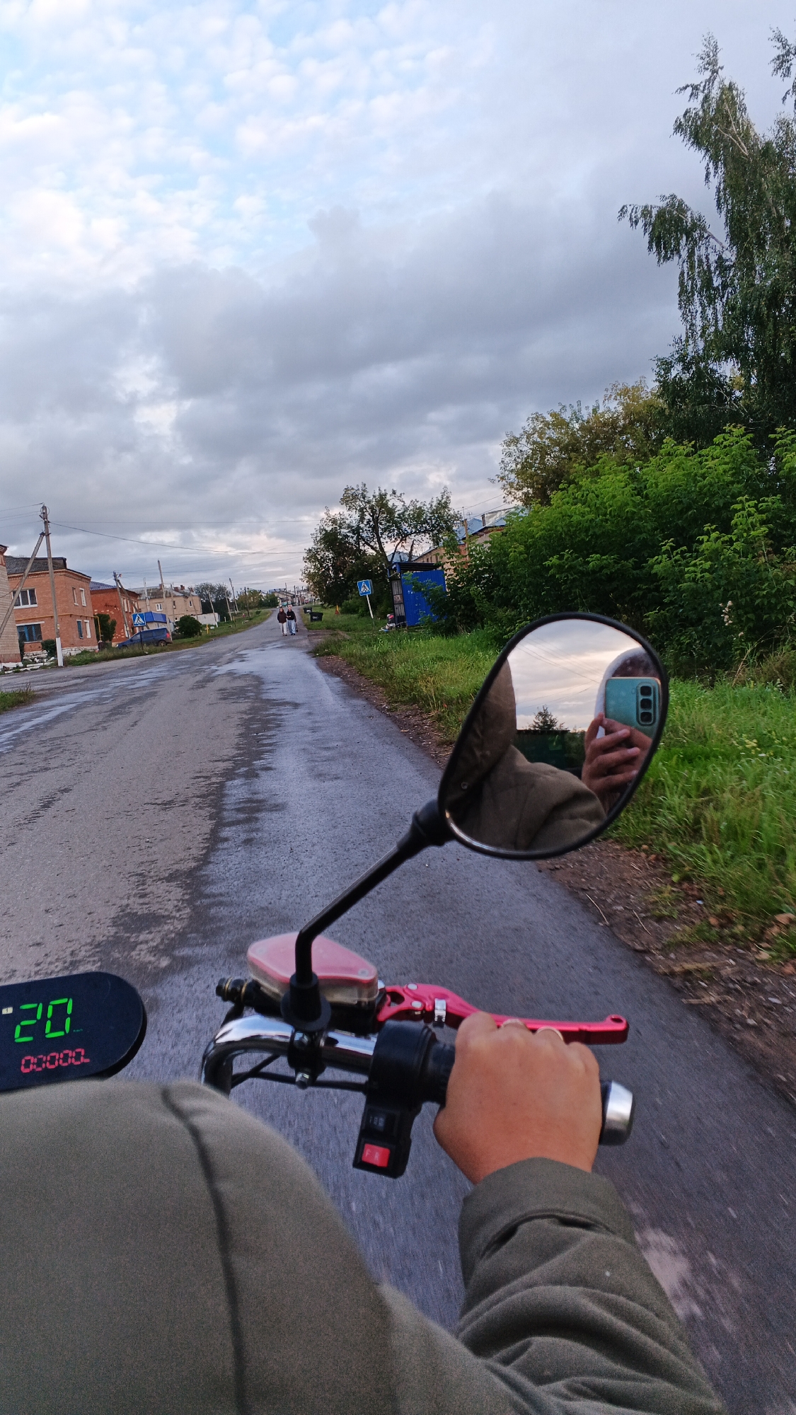 The image captures a first-person perspective from a person riding a scooter on a wet asphalt road in the village of Podgornoye, Russia. The rider's hand is visible, gripping the handlebars, which are adorned with a red brake lever and a switch. A digital display on the scooter shows "20" in green digits and "00000" in red. In the rearview mirror, the rider's reflection is visible, holding a smartphone with a blue case. The road is lined with green grass and trees on the right side. On the left side of the road, there are houses with brick facades and one with a white exterior. The sky is overcast with clouds. Two figures are walking in the distance on the left side of the road.