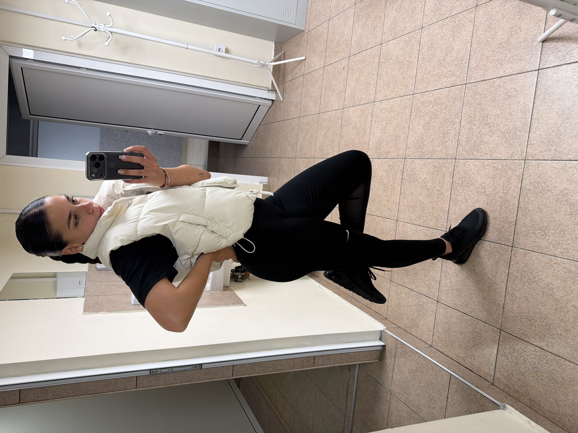 A young woman is captured in a full-body mirror selfie, standing with her right hand on her hip and holding an iPhone with a dark case in her left hand, which is raised to take the picture. She is wearing a cream-colored puffer vest over a black short-sleeved top, black leggings with a faint white Nike swoosh on the left thigh, and black athletic shoes (likely Nike Air Max style). Her dark hair is pulled back, and she wears a silver bracelet on her right wrist, with a small, stylized "E" tattoo visible on her right forearm. She has a neutral expression as she looks towards the phone.
The setting appears to be an indoor hallway or waiting area, possibly in Gabrovo, Bulgaria. The floor is covered with light brown/beige speckled square tiles. The walls are a plain light cream color. In the background, there is a white interior door with a dark or frosted glass panel, and a white, four-pronged coat rack is mounted on the wall to the left. A low ledge or counter, possibly part of the mirror's frame, is visible along the bottom edge of the image. The scene is brightly lit by indoor lighting.