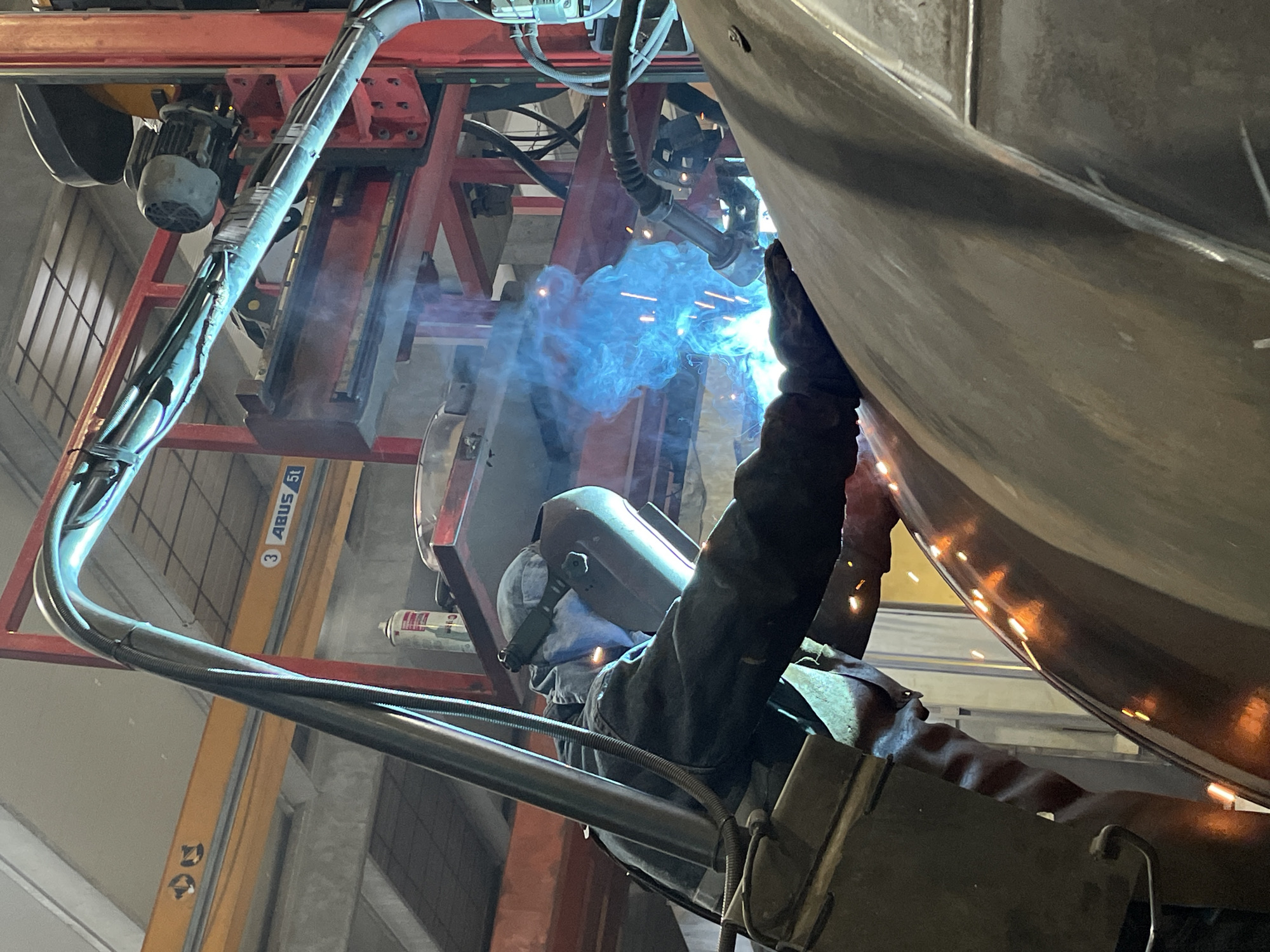 A welder is shown in Čeluga, Montenegro, welding a large, rounded metal object. The welder is wearing protective gear, including a welding helmet and gloves. Sparks and blue light are emitted from the welding point. The welder is positioned beneath the metal object, which appears to be suspended or held in place by a red metal frame and ABUS crane. The environment is an industrial setting with yellow-painted metal structures visible in the background.