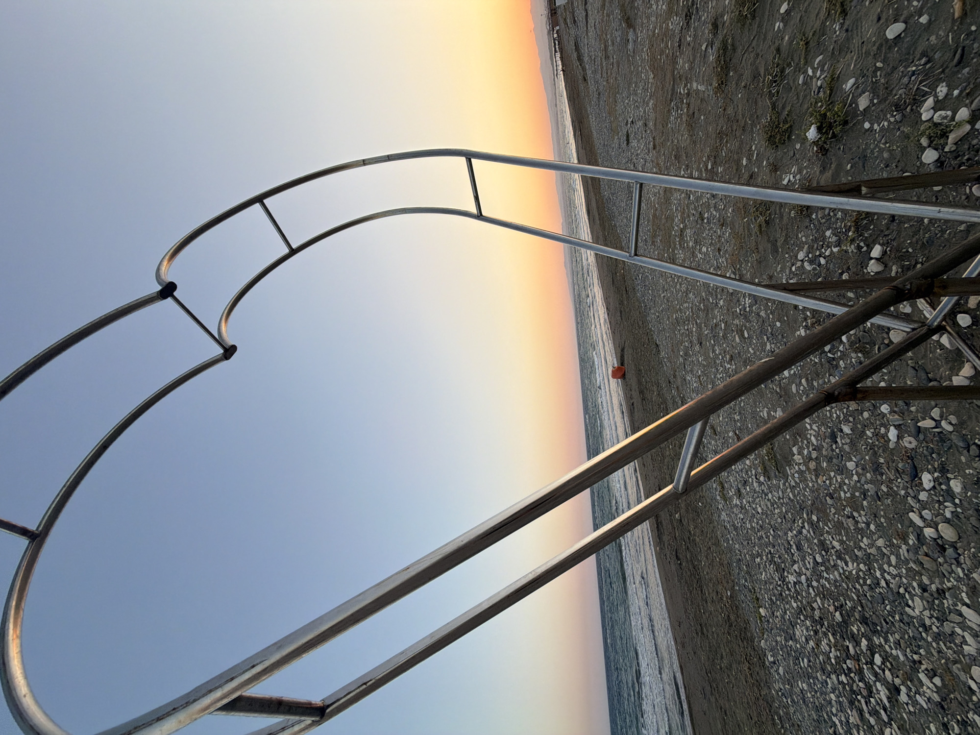 An outdoor, low-angle, wide shot captures a metal heart-shaped archway on a pebble beach at sunset in Meneou, Cyprus. The archway, constructed from polished metal tubing, stands prominently in the foreground, with its curved structure framing the horizon. The beach is composed of dark sand and smooth, multi-colored pebbles. Gentle waves of the sea create a soft line along the shore, and the water reflects the warm hues of the setting sun. The sky transitions from a clear, light blue near the top to shades of orange and yellow near the horizon, where distant mountains are faintly visible. A single, small orange buoy sits on the beach in the middle ground. The overall atmosphere is serene, with the vibrant sunset contrasting with the cooler tones of the beach and sea.