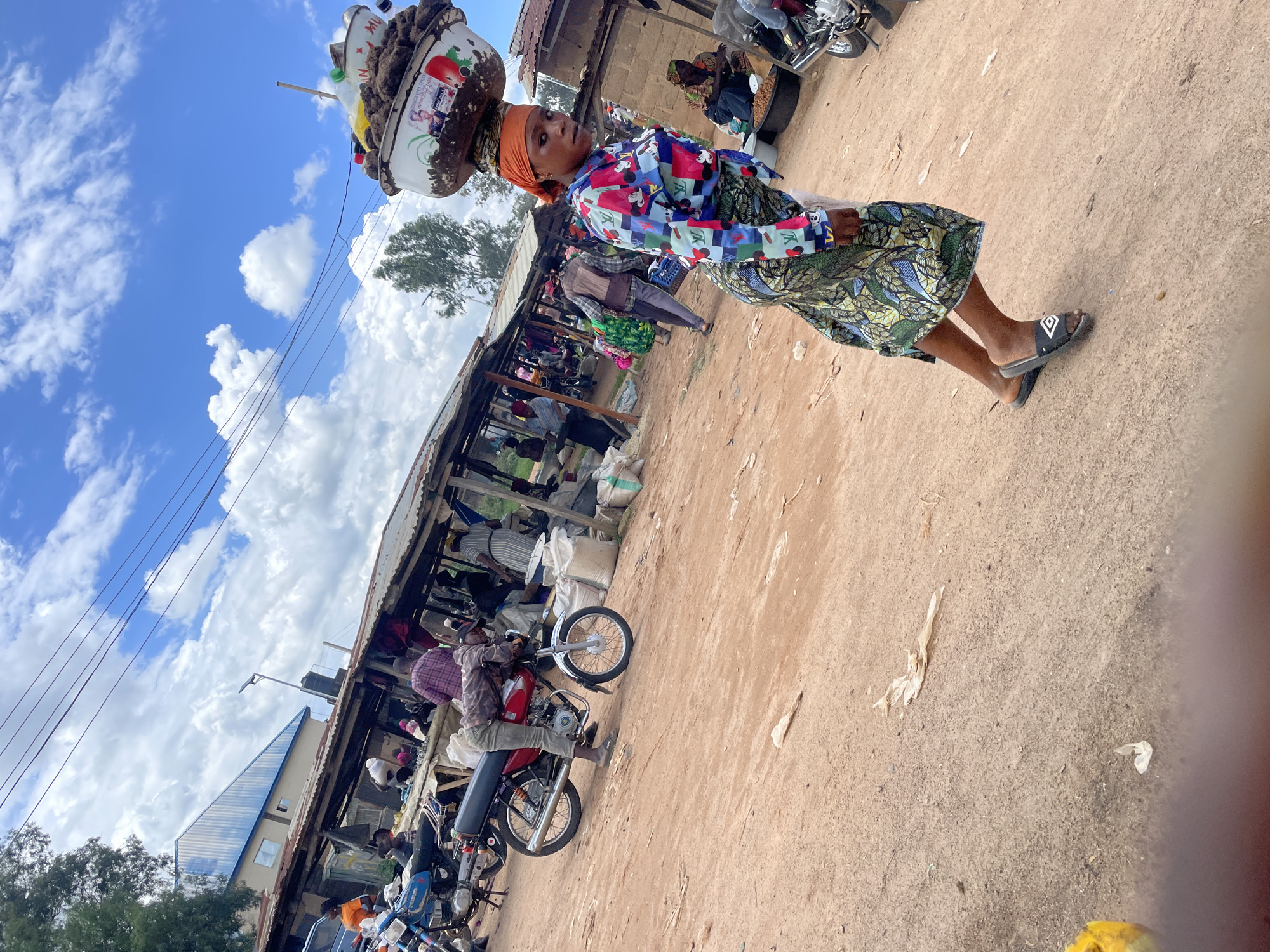 A medium shot captures a lively outdoor market scene in Wang, Nigeria, under a bright blue sky dotted with fluffy white clouds. In the foreground, a woman walks from right to left, balancing a large, overflowing bowl of goods on her head. She wears a vibrantly colored, long-sleeved shirt with a Mickey Mouse and LV pattern, and a green and gold patterned skirt. Her orange headwrap and black slide sandals complete her attire. The ground is a dusty, reddish-brown earth. Behind her, various market stalls with corrugated metal roofs are visible, with people engaged in commerce. Several motorcycles are parked throughout the market. In the background, a blue and white building stands under the expansive sky. The atmosphere suggests a busy market day.