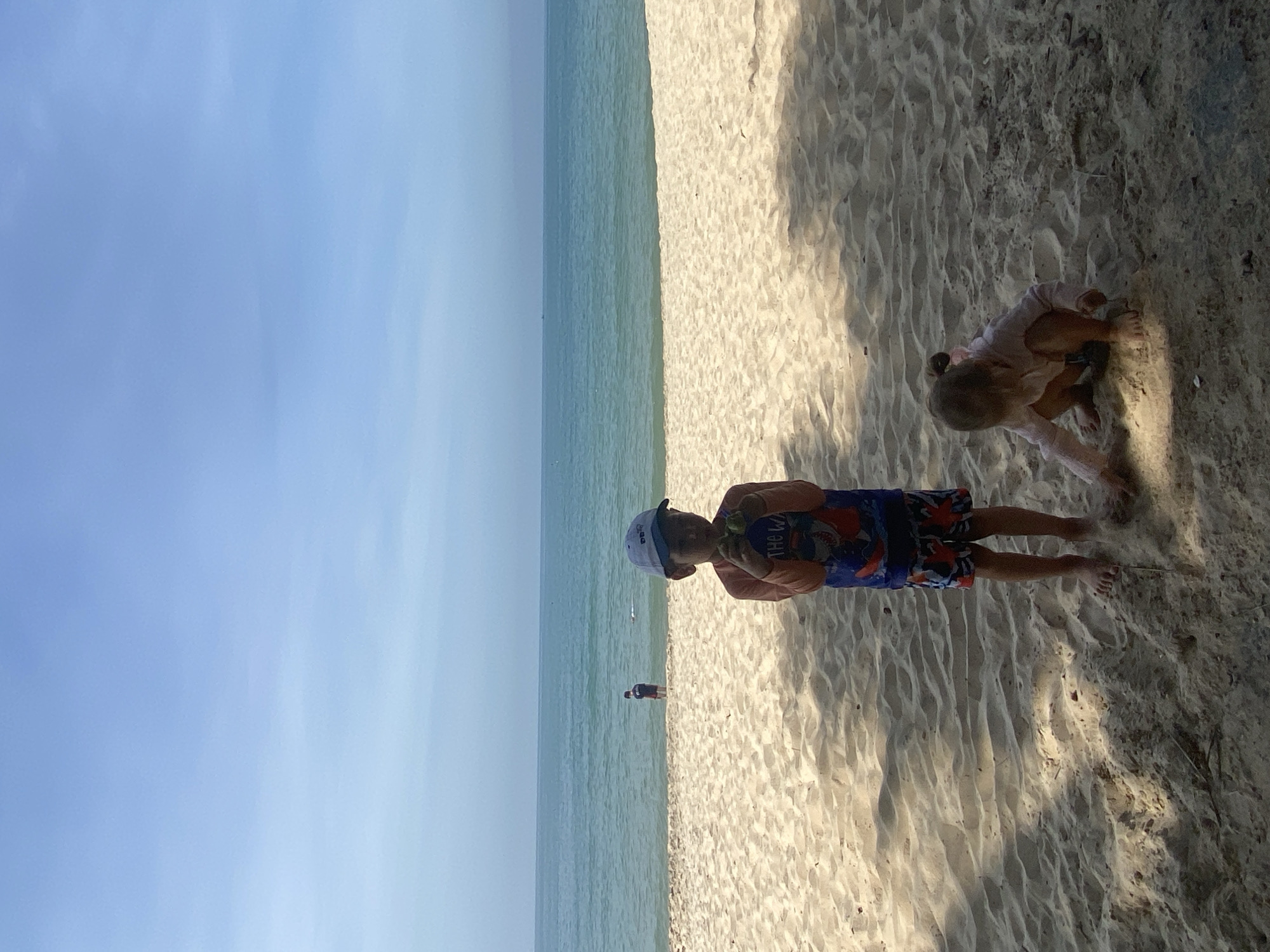 The image depicts a sunny day at a sandy beach in Phu Thuy Ward, Vietnam. The scene is divided into three main parts: a clear blue sky, a calm turquoise ocean, and a sandy beach. Two children are the primary subjects. In the foreground on the right, a young child wearing a pink shirt and dark shorts is crouched down, seemingly playing in the sand. Further into the frame, to the left, a young boy wearing a white cap and colorful shark-patterned swim trunks stands holding a green object. He is looking down, possibly at the object in his hands. In the distance, two other figures are visible near the water's edge. The lighting suggests it is either late morning or early afternoon, with the sun casting shadows on the sand. The overall atmosphere appears peaceful and recreational.