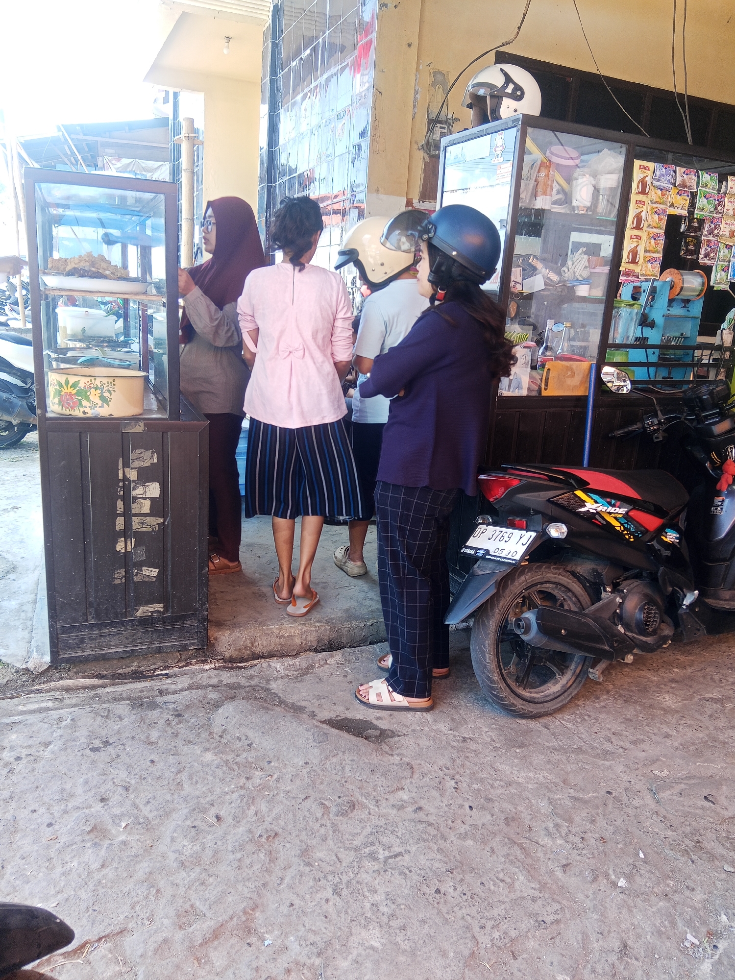 In front of a shop in Makale, Indonesia, four people are visible, two women and two men, with one of the men wearing a helmet. The woman on the left is wearing a dark headscarf and glasses, and she is looking at something in a glass display case. The woman next to her is wearing a pink shirt and a striped skirt, and she is looking away from the camera. The third person from the left, a man, is wearing a white helmet and a light-colored shirt. The fourth person, a man, is wearing a blue helmet and a dark jacket. A motorcycle is parked to the right of the group. The ground is concrete, and there are some plants visible in the background. The shop appears to be selling snacks or other small items.