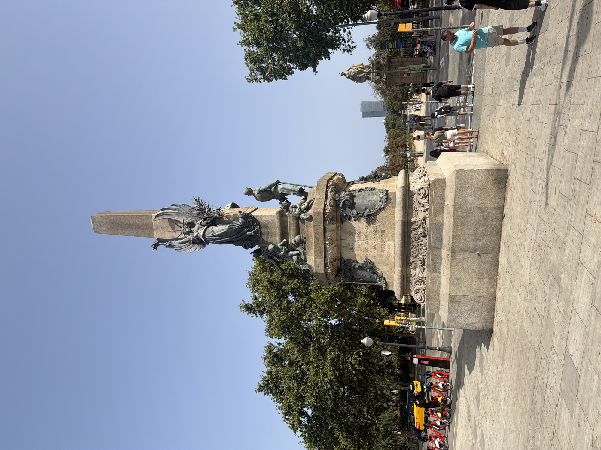 The image shows a monument in Barcelona, Spain, likely the Monument a Rius i Taulet. The monument is a large stone obelisk topped with a bronze sculpture of a winged figure. At the base of the monument are various bronze figures, including a standing male figure and a child. There are inscriptions on the monument that are partially visible.
The scene is outdoors on a sunny day with a clear blue sky. The monument is situated in a plaza paved with stone tiles. To the left of the monument, there is a row of red and white rental bicycles. A yellow taxi is parked behind the bicycles, and a person in dark clothing is standing near the bicycles.
In the background, there are trees, streetlights, traffic lights, and several people walking. Across the street, there is another monument or statue with a golden figure on a pedestal. In the far distance, a tall modern building can be seen. The people in the image are casually dressed, suggesting a public area or tourist attraction. Some people are walking, while others are standing and looking around. One man in a light blue polo shirt and beige shorts is standing in the foreground, looking at his phone.