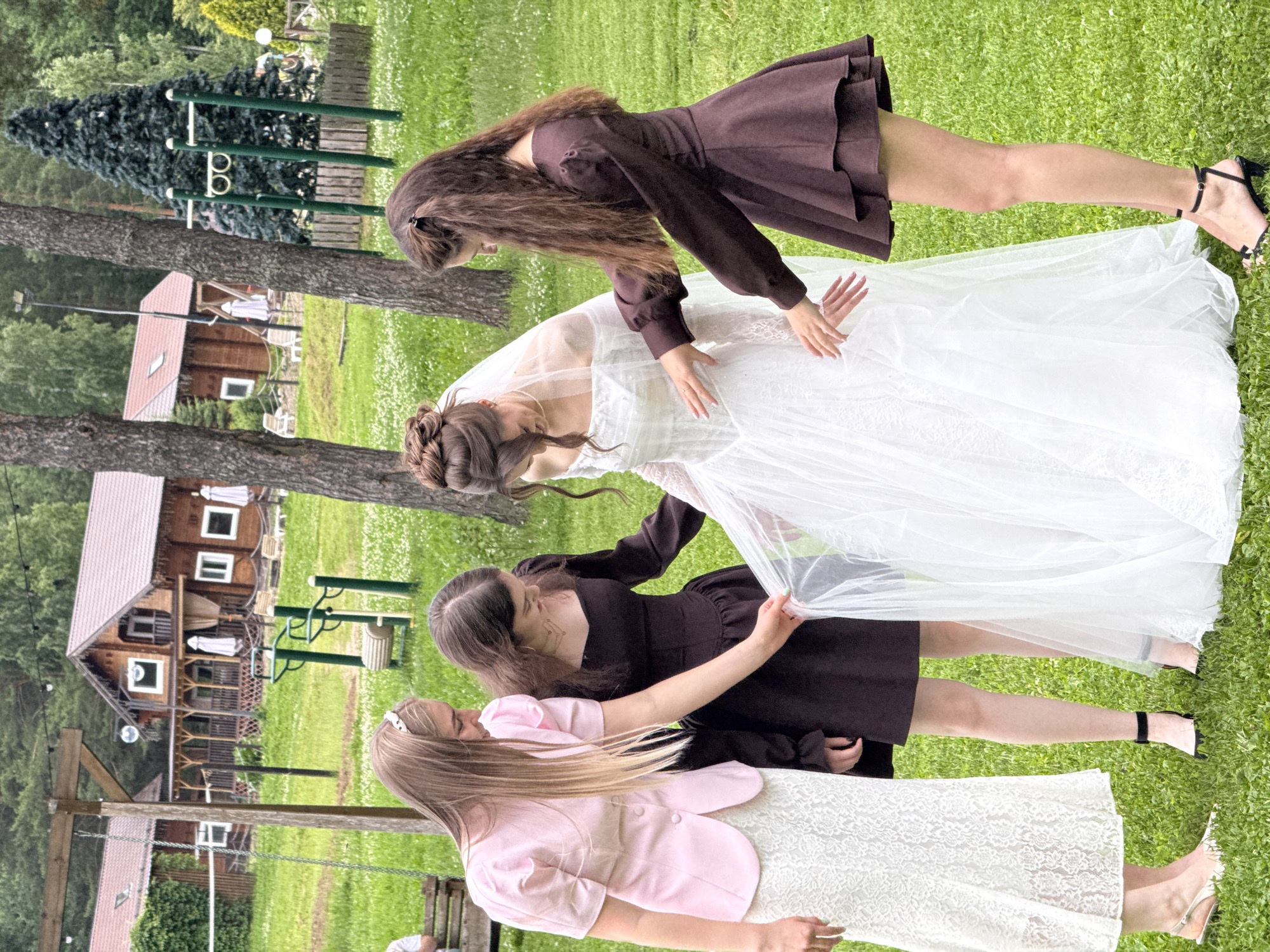 Four young women are gathered in a grassy outdoor area, with a wedding scene evident. In the center, a bride in a white wedding dress, complete with a veil, is being attended to by three other women. One woman to the bride's left, wearing a pink jacket over a white lace skirt, appears to be adjusting the bride's veil or dress. To the bride's right, two women in dark brown dresses are also interacting with the bride, one touching the bride's dress and the other with her hands outstretched. The background features wooden buildings with red roofs, trees, and outdoor recreational equipment, suggesting a rural or park-like setting. The overall scene captures a moment of preparation or adjustment before a wedding ceremony.