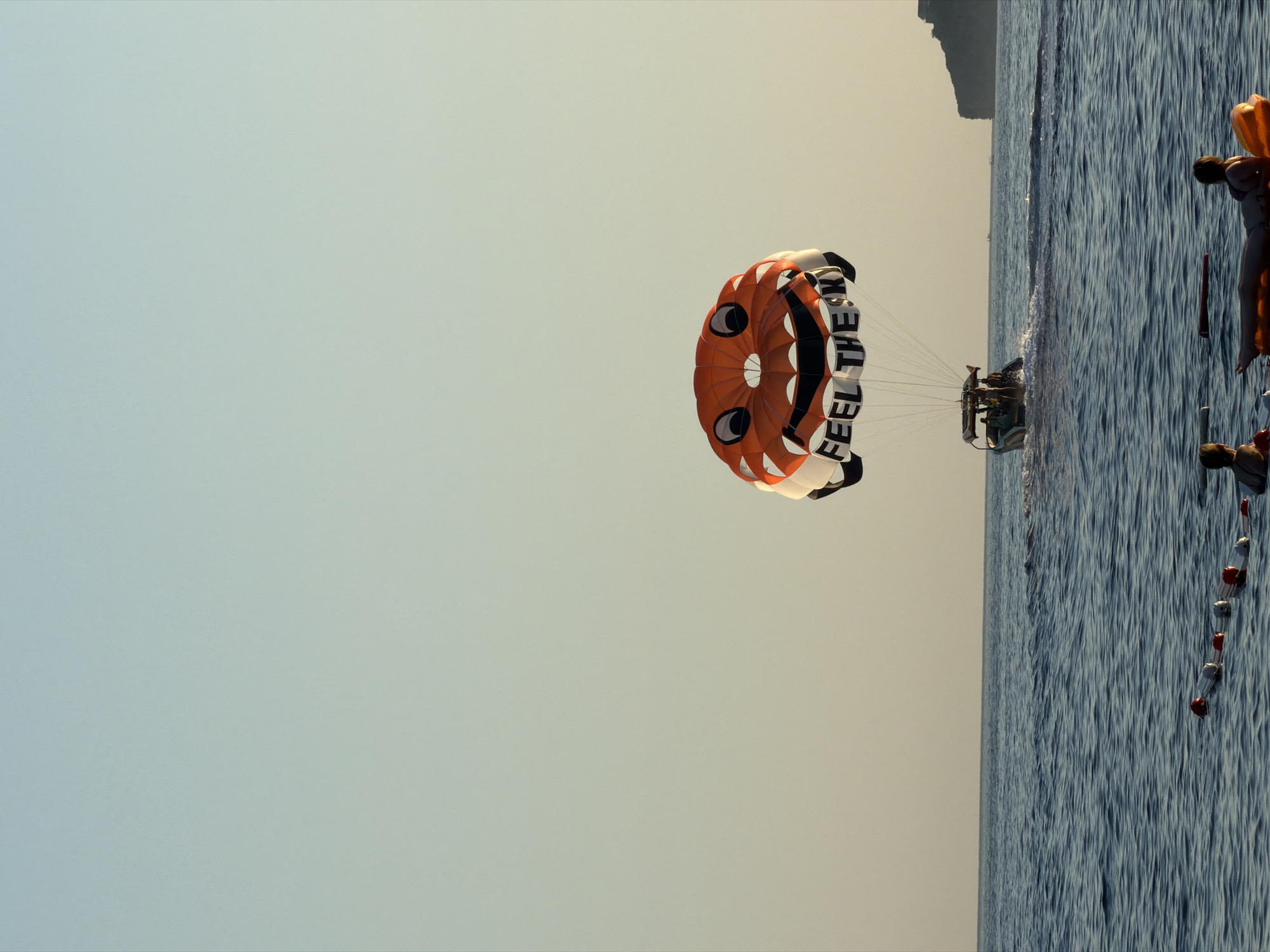In the city of Boreti, Montenegro, a parasail adorned with a smiley face is visible against a clear sky. The parasail, which displays the words "FEEL THE SKY," is towed by a boat carrying several passengers. The boat is creating a wake in the calm, blue water. In the foreground, two individuals are partially submerged in the water, with one person relaxing on an inflatable. A safety line with buoys stretches across the water in the mid-ground. The horizon shows a rocky coastline on the right side of the image.