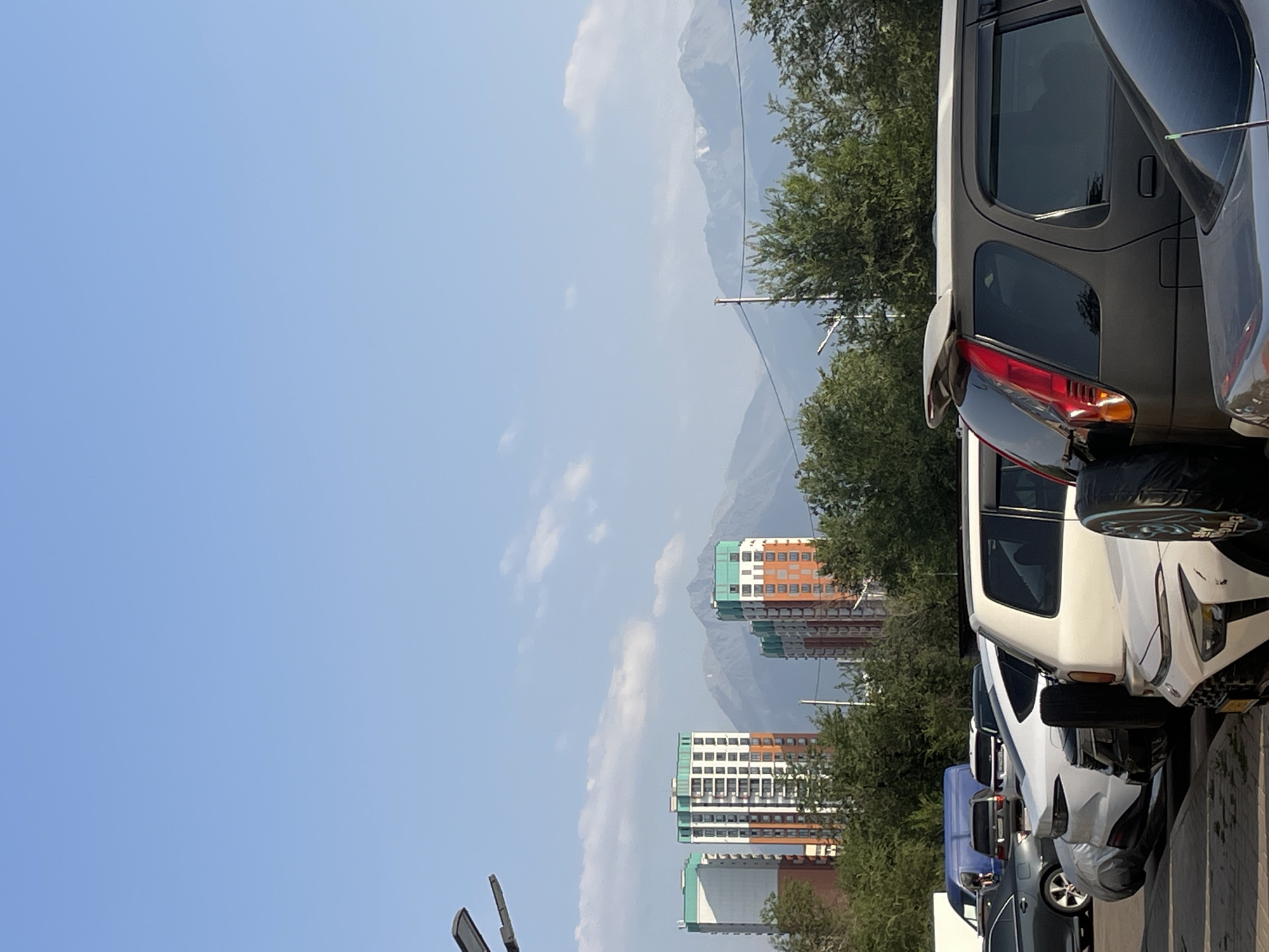 The image shows a daytime outdoor scene in Almaty, Kazakhstan, with a clear blue sky and scattered clouds. In the background, several modern, multi-story buildings with orange, white, and green facades are visible, interspersed with lush green trees. Beyond the buildings, snow-capped mountains rise majestically against the horizon, suggesting a location with proximity to natural landscapes. In the foreground, a row of parked cars, including SUVs and sedans, are lined up, some partially covered. The overall environment appears to be a residential or urban area with a backdrop of natural beauty.
