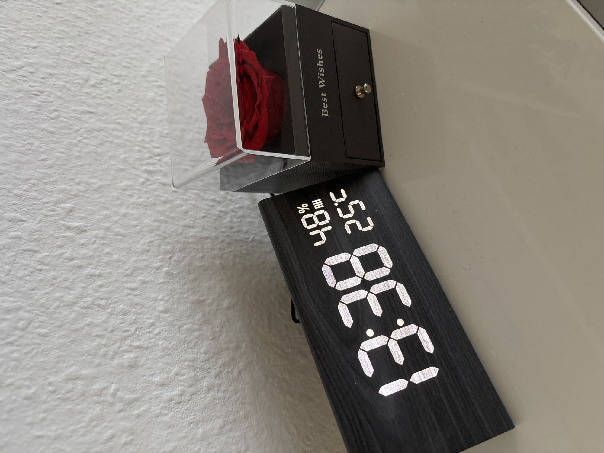 The image presents an indoor scene with two primary objects situated on a light-colored surface, positioned against a textured white wall. On the left, a digital clock with a black, wood-grain finish displays white LED segments showing '13:38' (1:38 PM), '48% RH' (relative humidity), and '25°C' (temperature). To the right of the clock, a black rectangular box contains a single red preserved rose, visible through a transparent acrylic lid. The front of the box is inscribed with the text "Best Wishes" and includes a small drawer with a silver-colored knob. The textured white wall forms the background in the upper half of the image, while the smooth, light beige surface on which the objects rest occupies the lower half. No individuals are present.