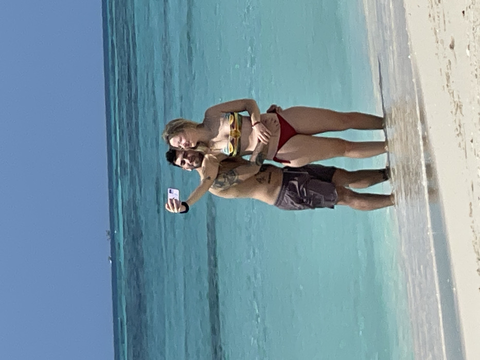Two individuals, an adult male and an adult female, are standing in shallow, clear turquoise water on a sandy beach in Zanzibar, Tanzania. The male, with dark hair, a beard, and visible tattoos on his right arm and torso, is wearing dark swim shorts. He holds a white smartphone extended in his right hand, positioned to take a selfie of himself and the female. His left arm is wrapped around the female's waist. The female, with blonde hair, is wearing a multi-colored strapless bikini top and red bikini bottoms. Her right hand rests on her abdomen, and her left hand is positioned on the male's lower back. The water reaches approximately ankle to calf-deep around their legs. The background consists of expansive turquoise ocean extending under a clear blue sky, with two small, indistinct boats visible on the distant horizon.