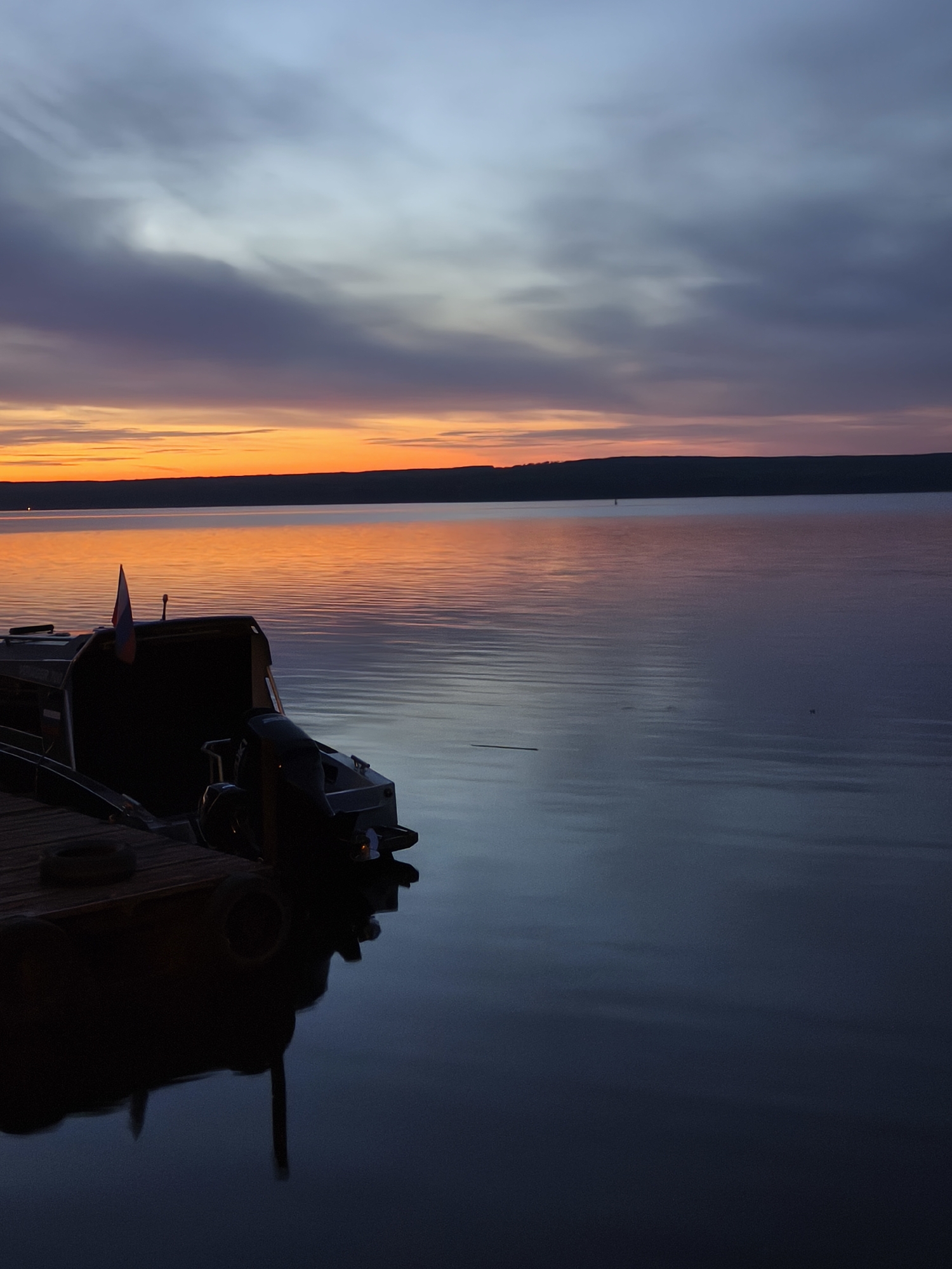 The image captures a serene sunset over the water in the city of Chaikovsky, Russia. The sky transitions from orange at the horizon to purple and grey higher up, reflecting in the calm water. A boat is docked at a wooden pier in the foreground, with a Russian flag visible on the boat. The water is mostly still, with gentle ripples on the surface. The background features a dark, low-lying land mass across the water.