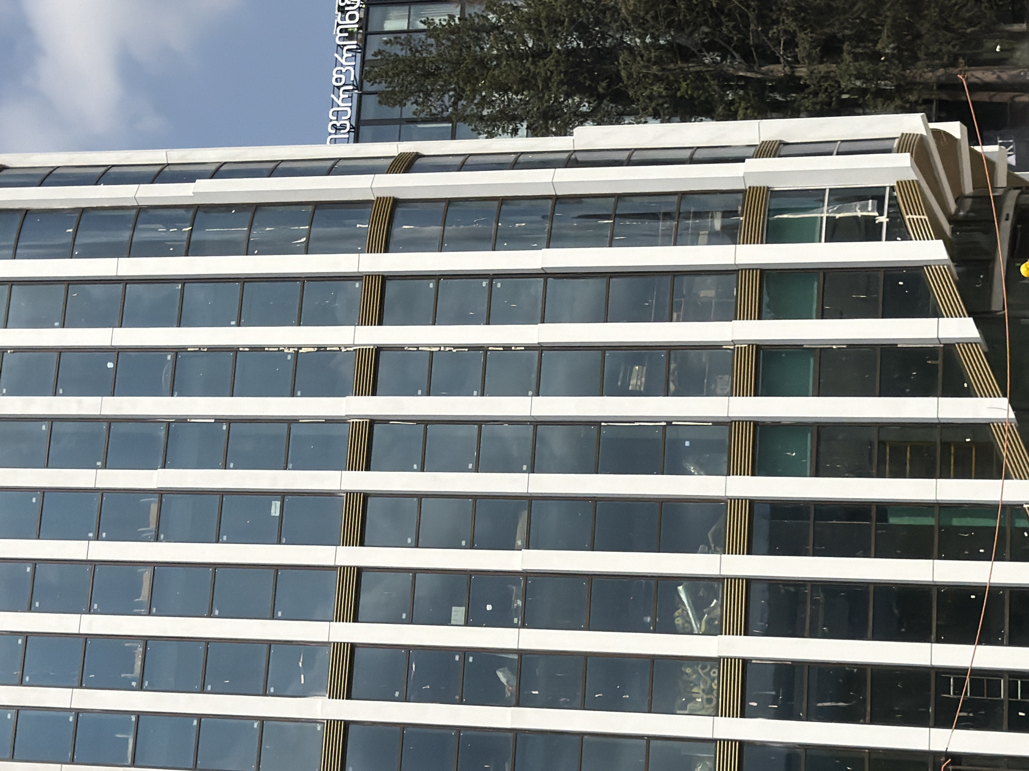 The image shows a modern glass-faced building with a curved facade and white vertical fins, set against a bright sky with scattered clouds. The windows appear to be dark tinted and reflect the sky. Some of the windows show interior contents, including what appears to be construction materials and possibly furniture. A neon sign in Georgian script is visible on an adjacent building to the right, partially obscured by a tall evergreen tree. The sign reads "ტბილისი მერია" which translates to "Tbilisi City Hall". The overall scene suggests an urban environment in Tbilisi, Georgia, possibly depicting a new or under-construction building in proximity to a government facility. The lighting suggests it is daytime.