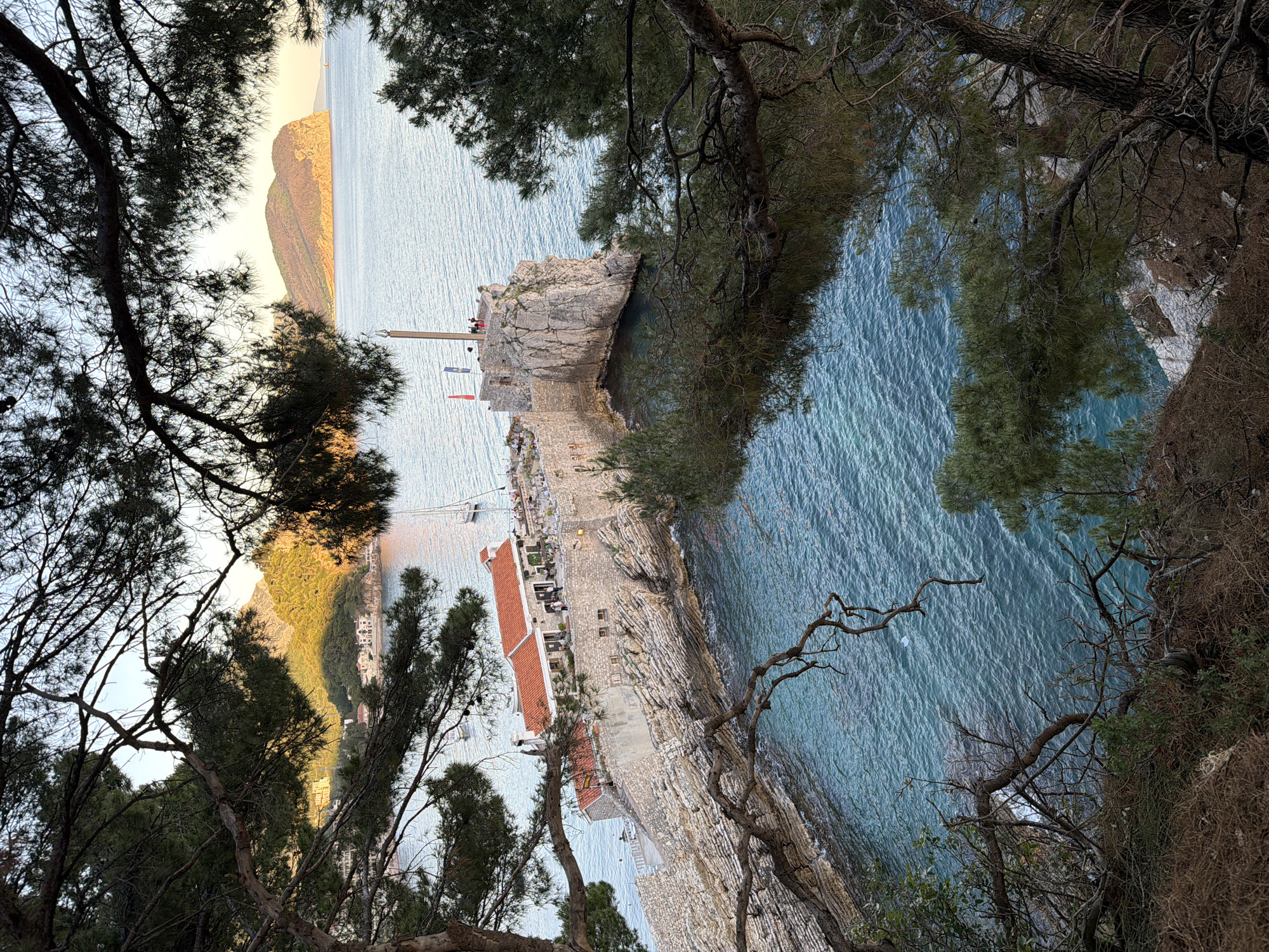The image captures a view of Petrovac centar, Montenegro, from an elevated perspective, possibly from a nearby hillside. The Adriatic Sea dominates the scene, with its turquoise water reflecting sunlight. A rocky peninsula extends into the sea, featuring a stone fortress with a red-roofed building and a flagpole. Trees frame the view from the sides, with dark branches contrasting against the bright sea and sky. A mountain range is visible in the distance, shrouded in a soft, warm light. The focus appears to be on the coastal architecture and the natural landscape.