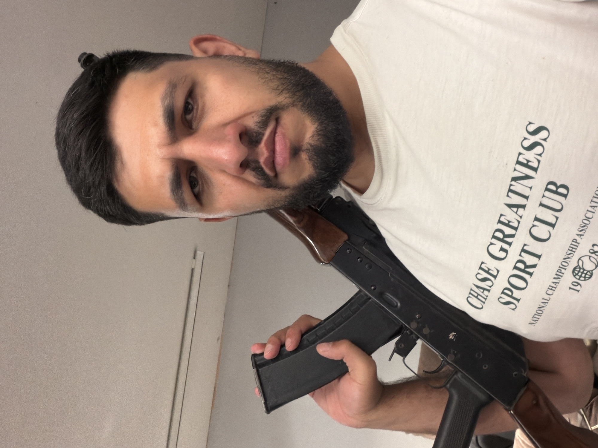 A man with short black hair and a beard is holding an AK-47 rifle. He is wearing a white t-shirt with "CHASE GREATNESS SPORT CLUB" printed on it. The location is not evident in the image, and there is no indication it is in Beryozovka, Russia. The environment appears to be indoors, with a white wall behind the man. He is holding the rifle's magazine with his left hand. The man is looking directly at the camera.