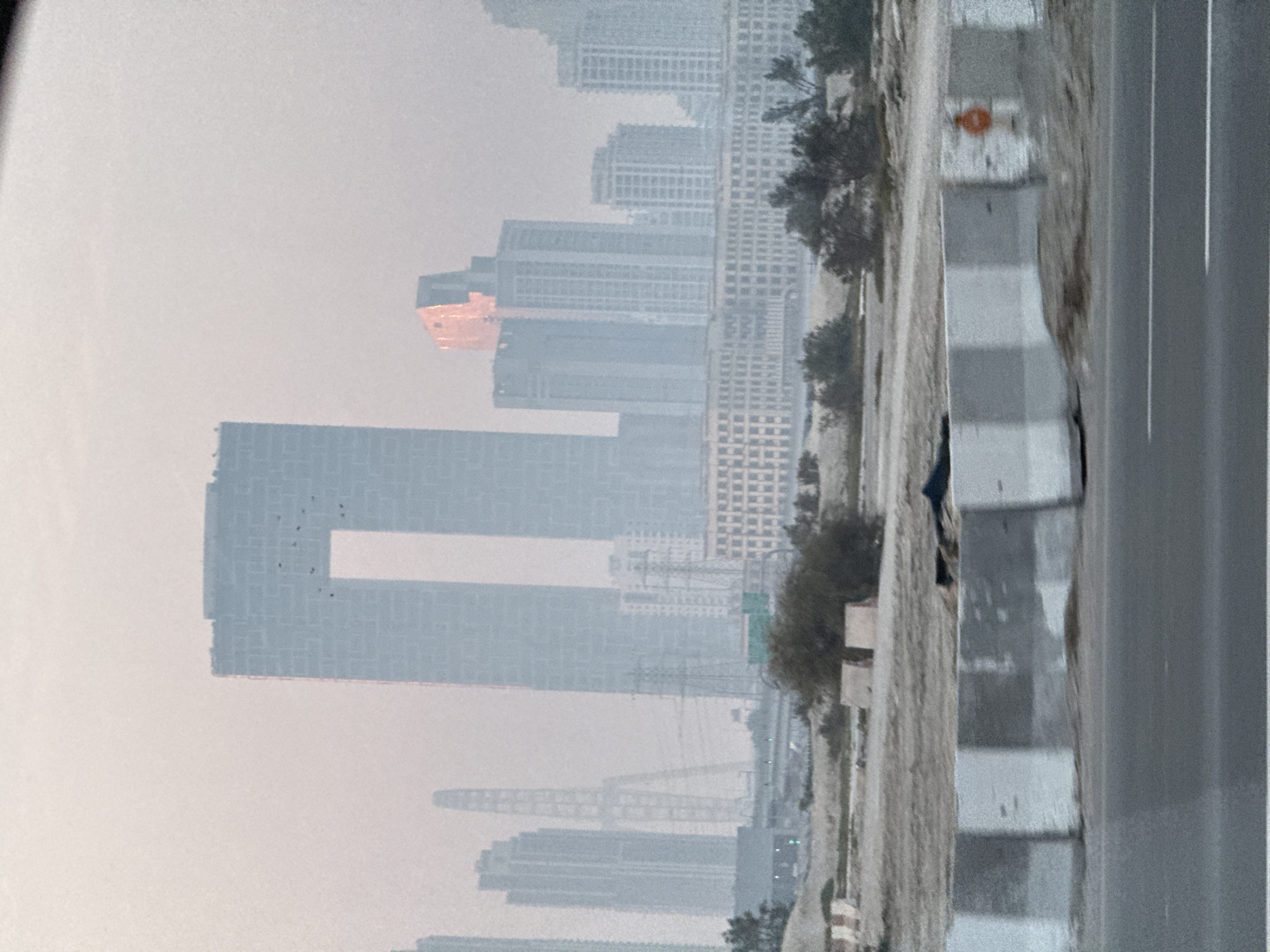 The image depicts a hazy cityscape in Dubai, United Arab Emirates. In the foreground, a grey asphalt road is visible with white lane markings. A barrier made of concrete blocks with alternating grey and white sections runs along the edge of the road. Beyond the barrier, a dry, sandy landscape with sparse vegetation and some construction materials is present.
The middle ground features a collection of buildings. A prominent, light-blue rectangular skyscraper with a large rectangular opening in the center dominates the left side of the skyline. To its right, several other modern skyscrapers of varying heights and designs are clustered together. One of these buildings reflects a reddish-orange light, possibly from the setting sun or an external light source. Further back, a long, multi-story building with numerous windows stretches horizontally across the frame. In the distance, power transmission towers and other structures can be faintly discerned through the haze. The sky is a pale, overcast grey, suggesting a hazy or dusty atmosphere. No people are visible in the scene.