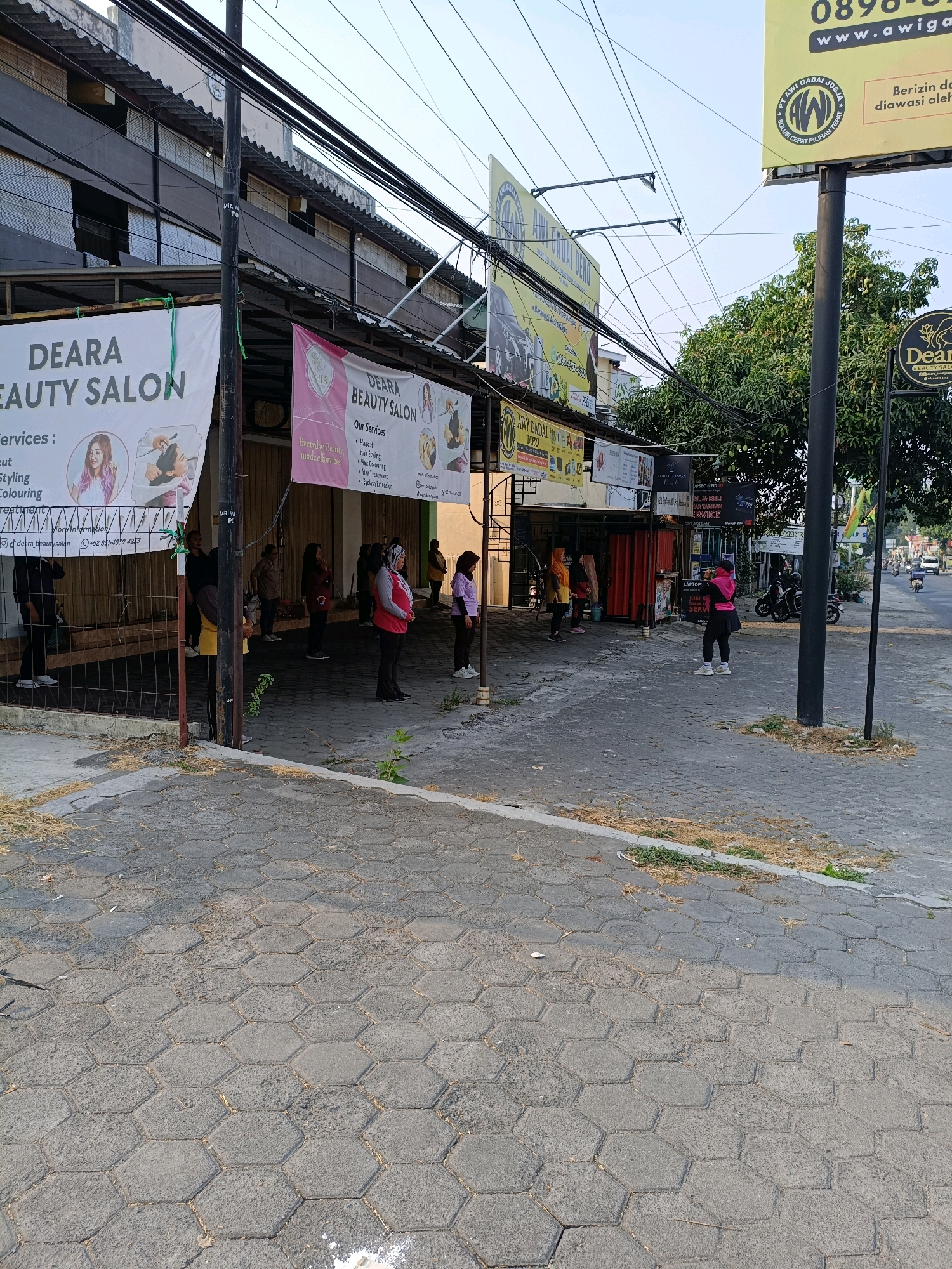 The image depicts a street scene in Ngemplak, Indonesia, likely in front of a row of businesses. The foreground features a textured gray hexagonal paver sidewalk. To the left, a white banner advertising "Deara Beauty Salon" with images of hair styling and coloring is partially visible. Further down the sidewalk, another banner for "Deara Beauty Salon" is present. A group of at least eight individuals, predominantly women, are standing in a line spaced apart, appearing to be engaged in some form of outdoor activity or waiting. They are dressed in casual clothing, with some wearing bright colors. Behind them, the storefronts are visible, with some featuring advertisements and signage, including a large yellow billboard with text and a logo. A utility pole with numerous electrical wires is prominent in the center of the frame. In the background, trees and a street with passing vehicles can be seen, suggesting a public urban environment.