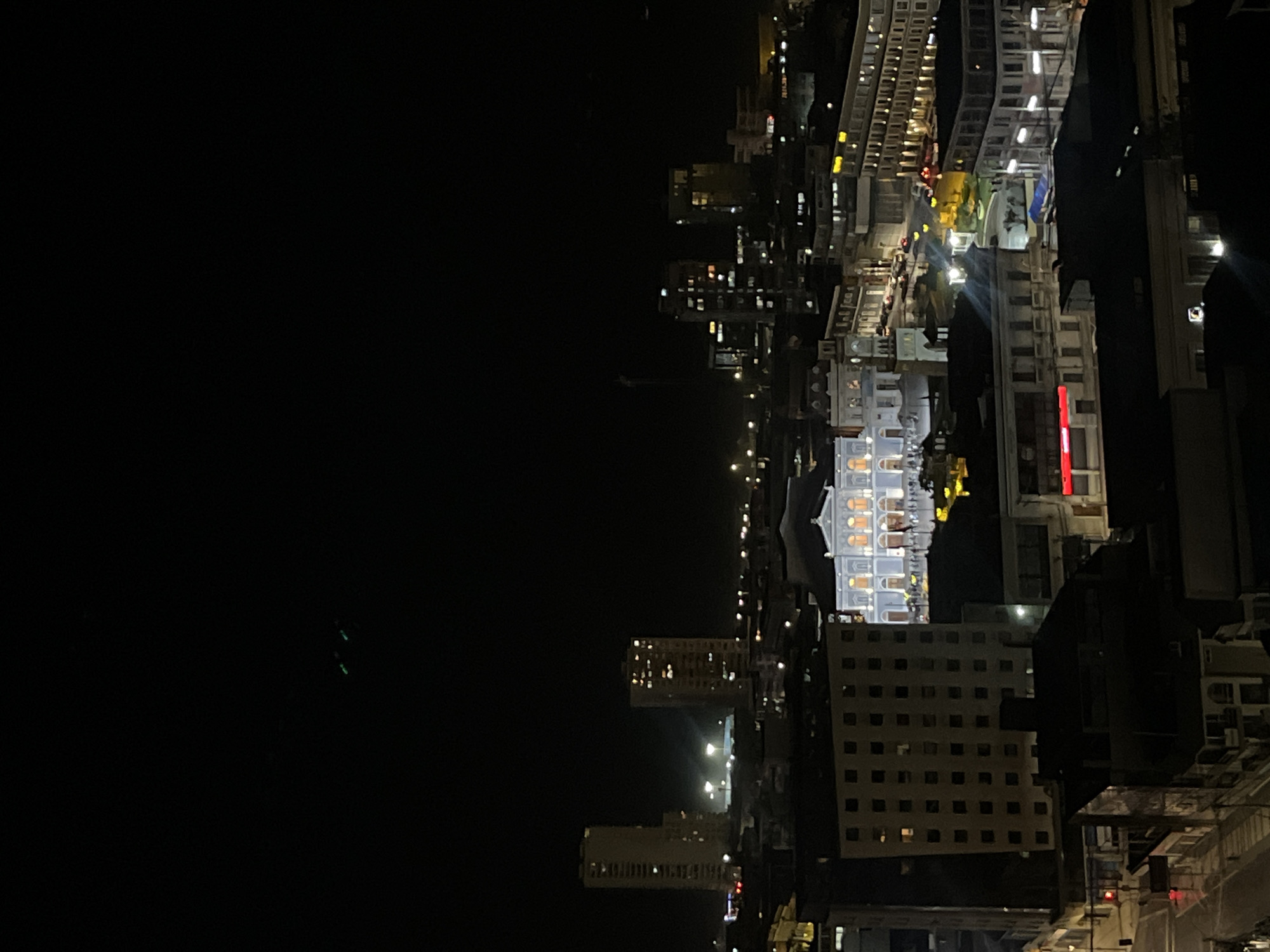 The image shows a nighttime view of a city, with illuminated buildings and streets. Several high-rise buildings are visible, along with a brightly lit, ornate structure that stands out from the surrounding buildings. Streetlights line the roadways, adding to the overall brightness of the urban landscape. The location is in the city of Unknown, Unknown.