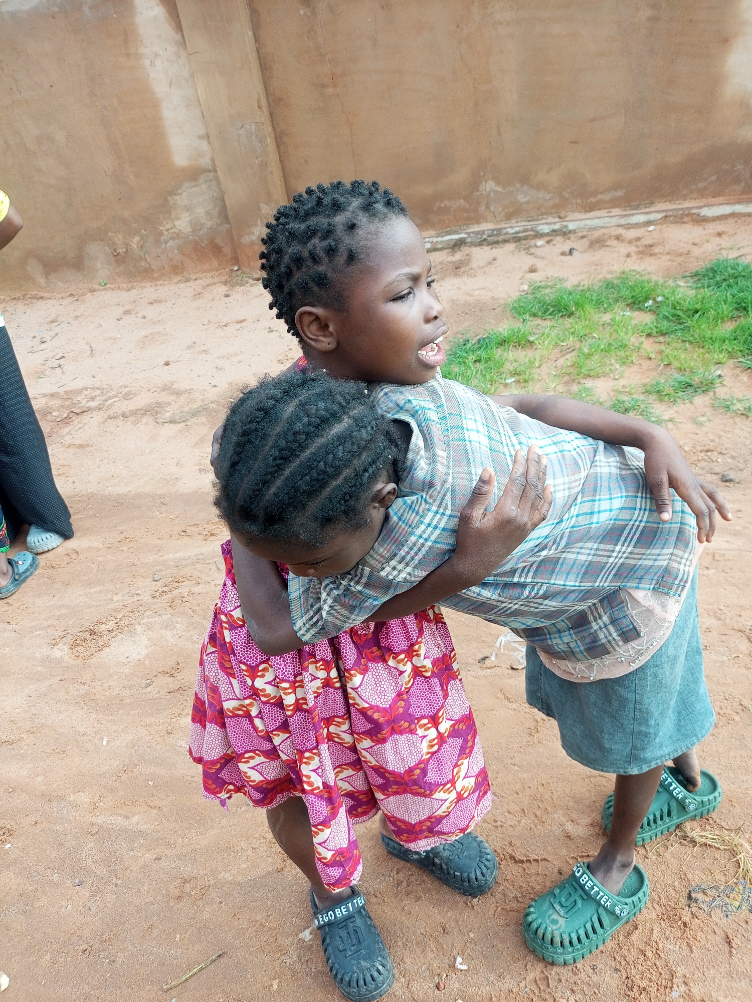Two young girls, appearing to be of Nigerian ethnicity and dressed in casual clothing, are embracing each other outdoors. One girl, facing away from the camera and looking to the right, wears a plaid shirt and a denim skirt. Her arms are wrapped around the other girl. The second girl, facing the camera and looking to the left, is wearing a colorful dress. Both are wearing green clogs. They are standing on sandy ground with patches of grass visible in the background, suggesting an outdoor environment. The wall behind them is a light brown, textured surface. The overall scene is simple and depicts a moment of interaction between the two children. The location is identified as Tempure, Nigeria.