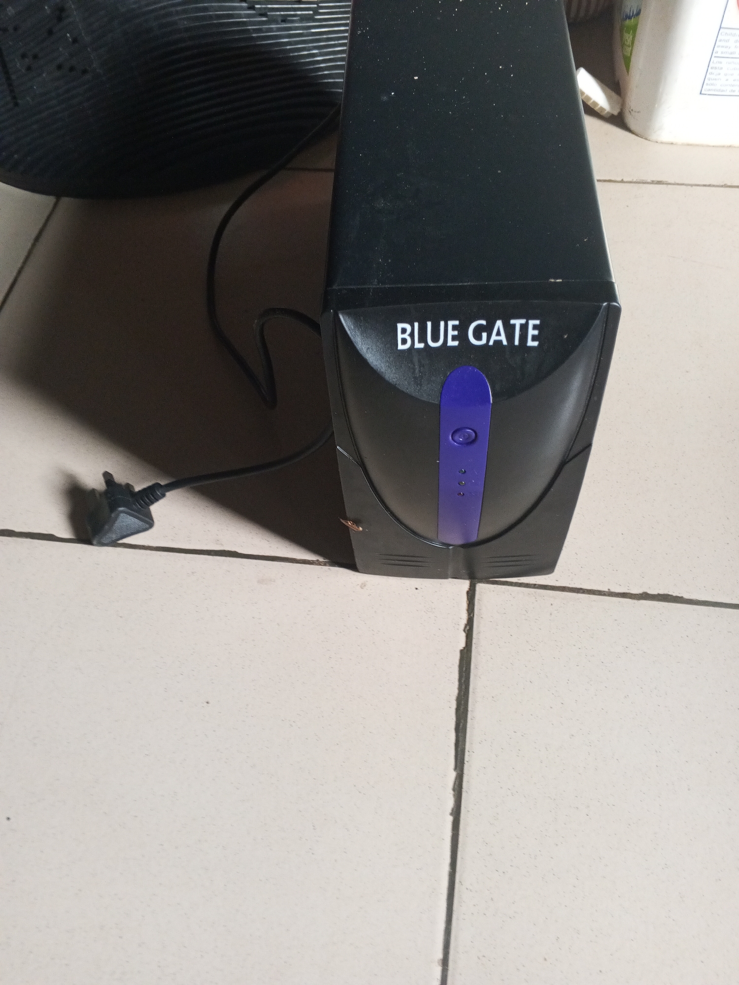 The image shows a black uniterruptible power supply (UPS) unit with "BLUE GATE" printed on it in white text. The UPS has a purple button on the front with three small indicator lights below it. A black power cord with a black plug is attached to the UPS and extends to the left, casting a shadow. The UPS is sitting on a tiled floor, with some dust visible. In the background, a white bottle is partially visible, and a black ribbed object, likely a computer monitor, is on the left side of the frame. The location is identified as Ashata, Nigeria.