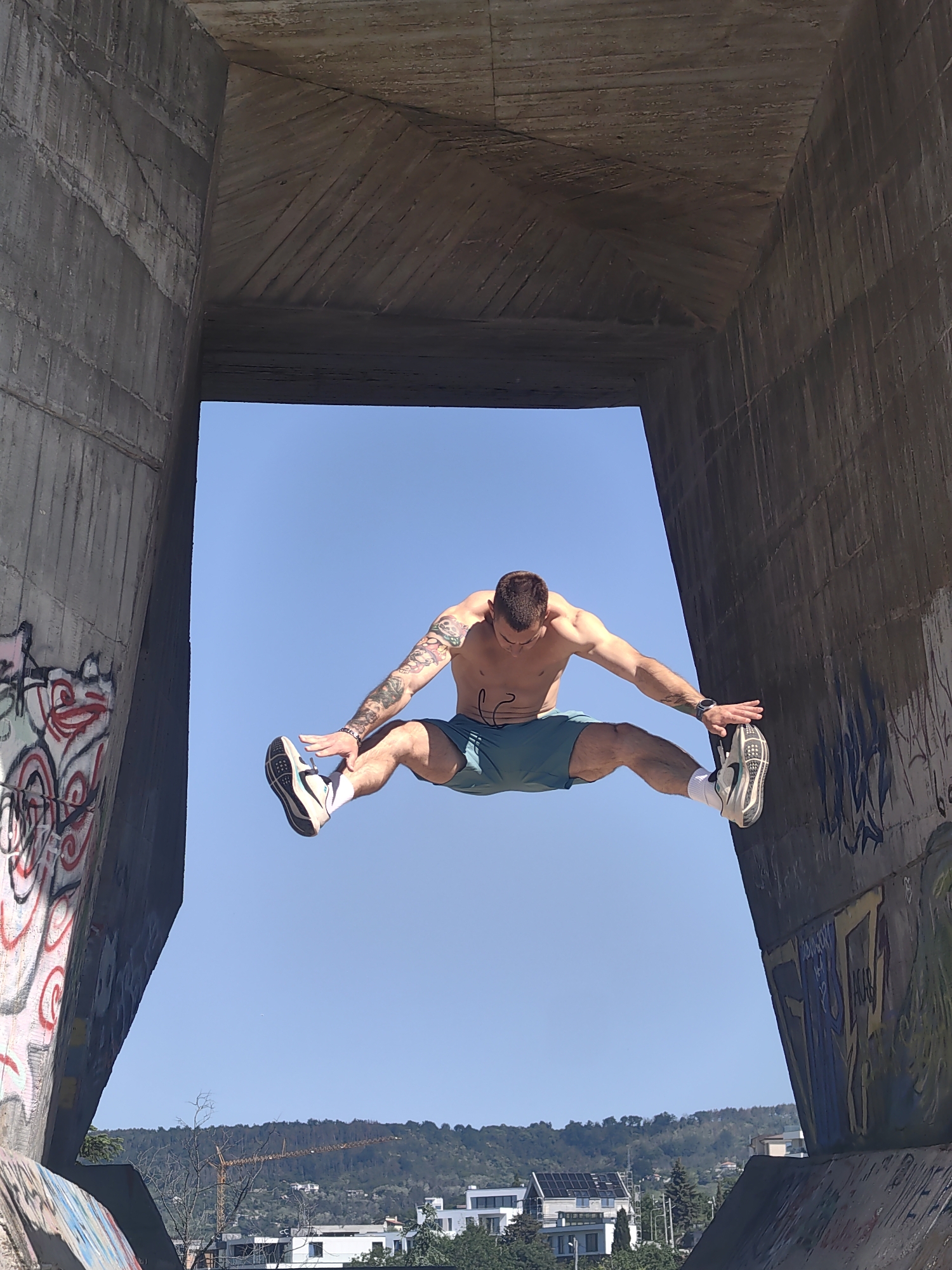 A man with tattoos on his arm is captured mid-jump, striking a wide, horizontal "V" shape with his legs and arms. He is wearing teal shorts, white socks, and sneakers. He is positioned in front of a concrete structure with an opening that frames the clear blue sky behind him. Graffiti is visible on the concrete structure. In the distance, a landscape with buildings, trees, and a hill is seen. A crane stands among the buildings. It appears this photo was taken in Varna, Bulgaria, based on the urban landscape and architectural features present in the background.