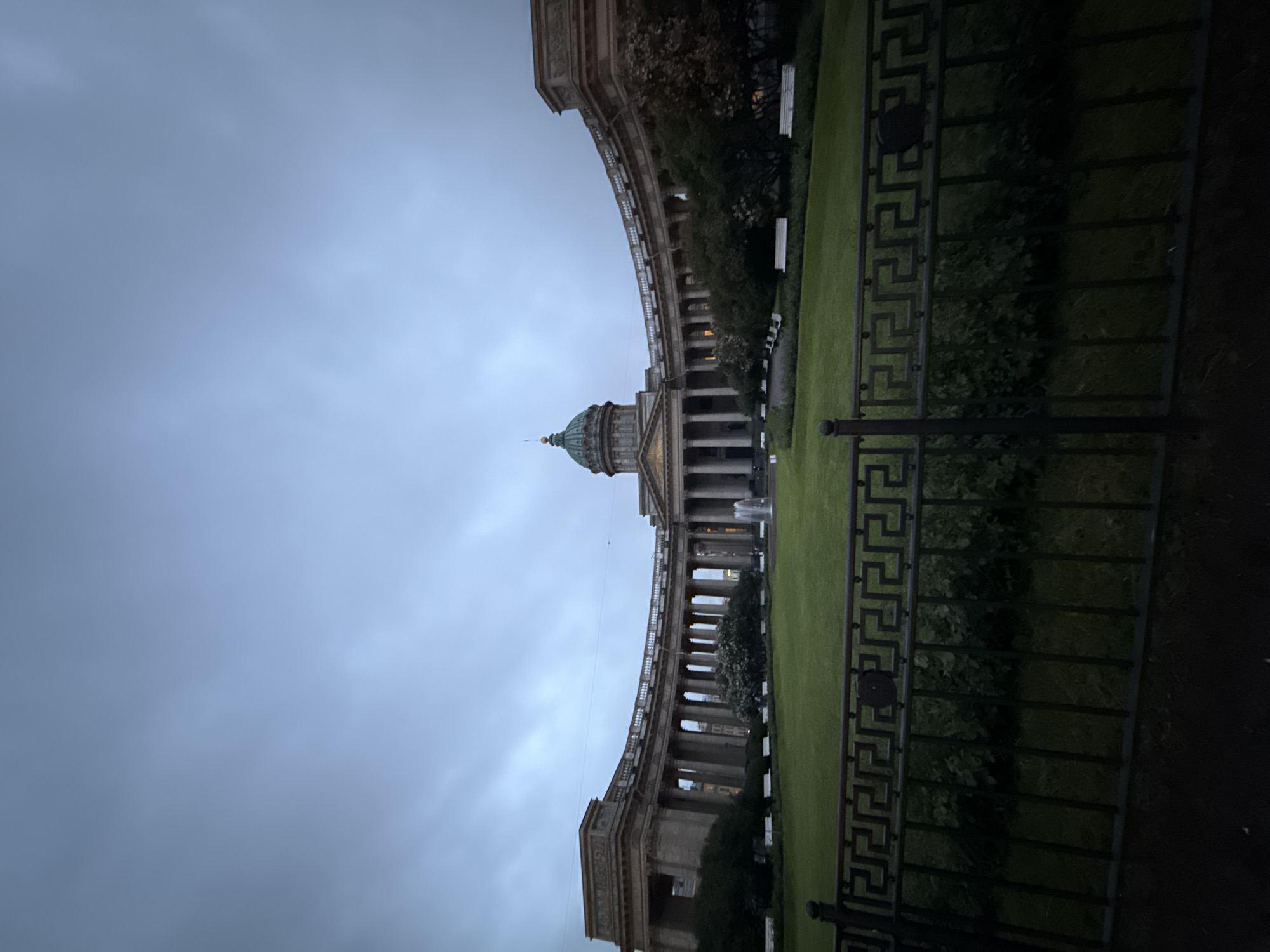 The image captures the Kazan Cathedral in Saint Petersburg, Russia, at dusk. The grand neoclassical structure, characterized by its semicircular colonnade and central dome, is illuminated by a dim, overcast sky. A well-maintained lawn stretches out in front of the cathedral, bordered by a decorative black metal fence in the foreground. White benches are visible on the lawn, suggesting a public space. A fountain is active in the middle ground, adding a touch of movement to the otherwise static scene. The overall atmosphere is serene and slightly melancholic due to the low light conditions and muted colors. There are no people visible in the frame.