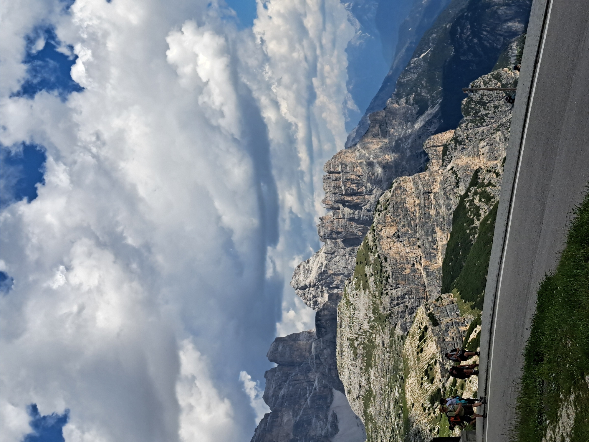 The image depicts a dramatic mountain landscape with a paved road in the foreground. The road curves along the edge of a steep, rocky cliff face that plunges down into a valley. The cliff is characterized by its layered rock formations, some of which are covered in sparse vegetation. The sky above is a mix of blue sky and dramatic, billowing clouds, suggesting a dynamic and potentially changing weather pattern.
Several people are visible walking on the road, some of them appearing to be a group of hikers or cyclists. They are dressed in casual outdoor clothing, and their presence suggests they are enjoying the scenery or perhaps on a journey through the mountains. The sunlight casting shadows on the road and cliffs indicates it is likely daytime, possibly late morning or afternoon.
The overall impression is one of grandeur and adventure, with the vastness of the mountains contrasting with the human presence on the road. The rugged terrain and cloudy sky create a sense of awe and perhaps a touch of wildness. There is no visible text in the image.