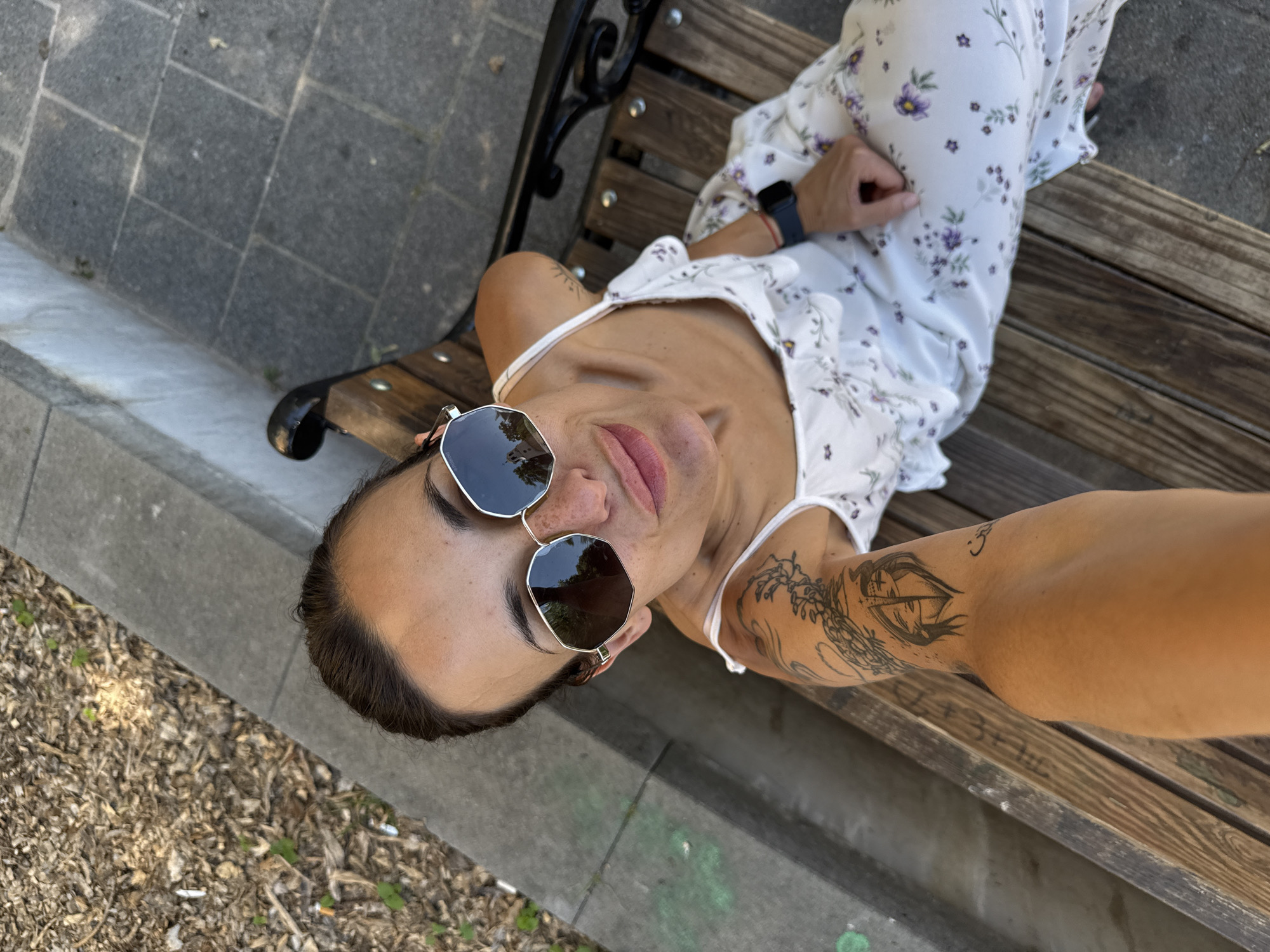 A person is sitting on a wooden park bench, taking a selfie. The person is wearing a white floral print dress and sunglasses. They have tattoos on their arm. The setting appears to be outdoors in a park, with a stone path and wood chips on the ground. The time of day is likely daytime, given the lighting. The person's expression is relaxed and casual. No visible text.