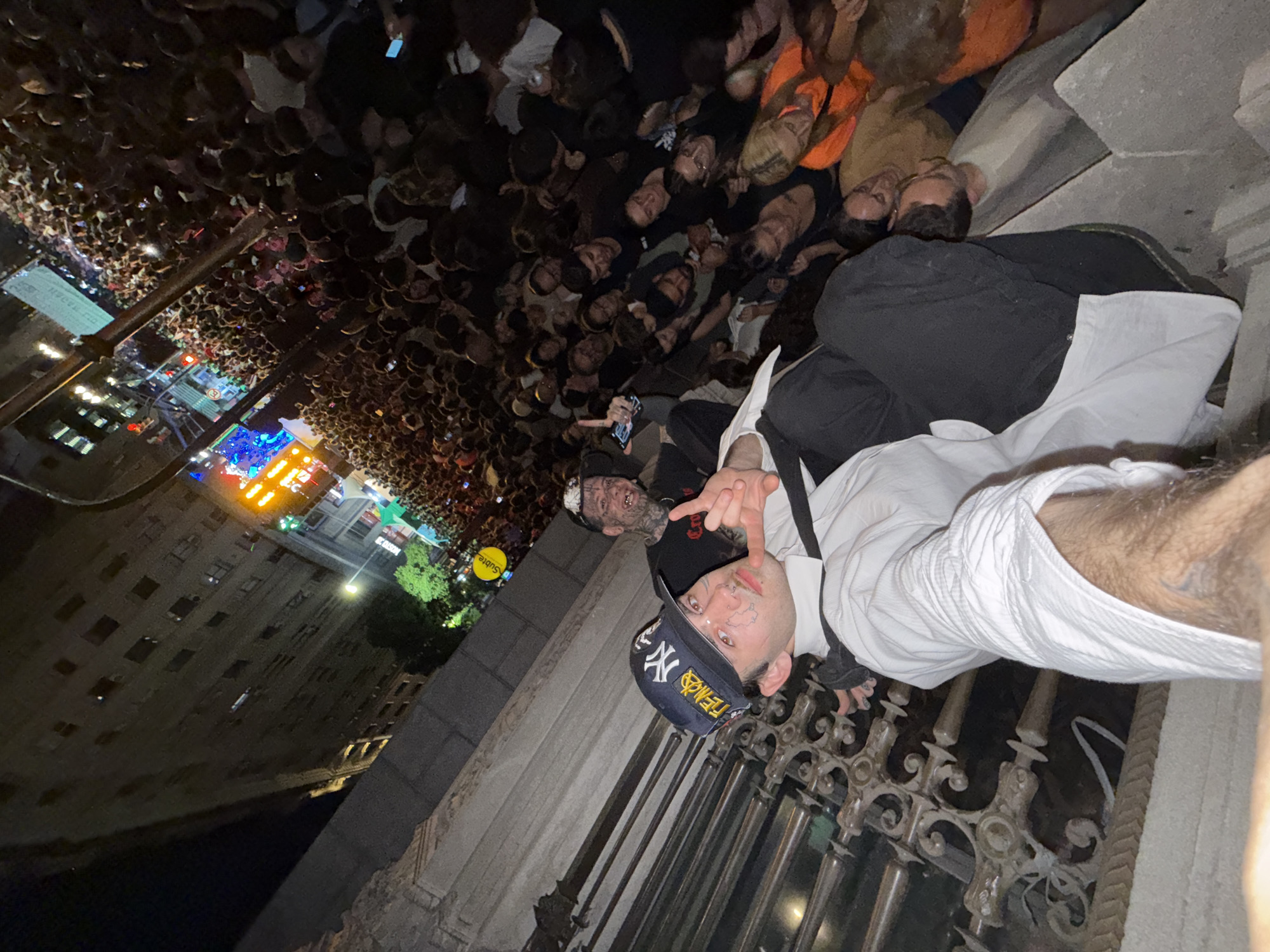 The image shows two men posing for a photo in front of a large crowd of people gathered at night. The man closest to the camera is wearing a New York Yankees baseball cap, a white shirt, and a black tie. He has tattoos on his face and is holding up a hand gesture. Behind him is another man with facial tattoos, wearing a black shirt. They appear to be on a balcony or elevated platform overlooking the crowd. Below them, the crowd fills the street, with buildings visible in the background. The location appears to be Buenos Aires, Argentina, based on the signs and architecture in the background. The lighting is a mix of streetlights and stage lights, suggesting a concert or event is taking place.