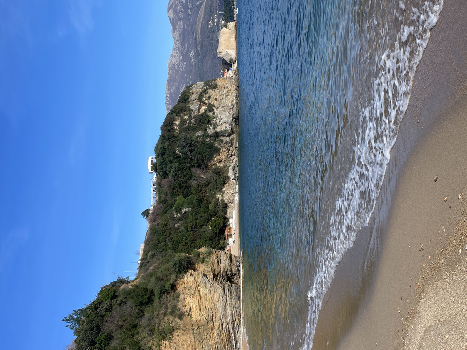 The image displays a coastal landscape in Budva, Montenegro, specifically Mogren Beach, observed from the sandy shore facing generally eastward. The foreground presents a light brown sand beach with scattered small pebbles, where calm, clear blue-green water forms gentle, white-foamed waves breaking at the water's edge. The midground consists of the Adriatic Sea, transitioning from lighter turquoise near the shore to deeper blue. To the left, a prominent, stratified rock cliff, composed of layered sedimentary rock, rises from the beach and is partially covered with dense green vegetation, including small evergreen trees and bushes. At the base of the cliff, on a smaller cove, several small structures with orange-brown roofs are visible. Further along the coastline to the right, the cliffs continue, heavily vegetated, with a light-colored, fortified structure or breakwater partially visible extending into the sea. In the background, atop the vegetated cliffs, white architectural elements are discernible, including a distinct white gazebo and parts of a building or terrace. Distant, rugged, grey-brown mountains appear under a clear, uniformly bright blue sky. No individuals, animals, or vehicles are present in the scene.