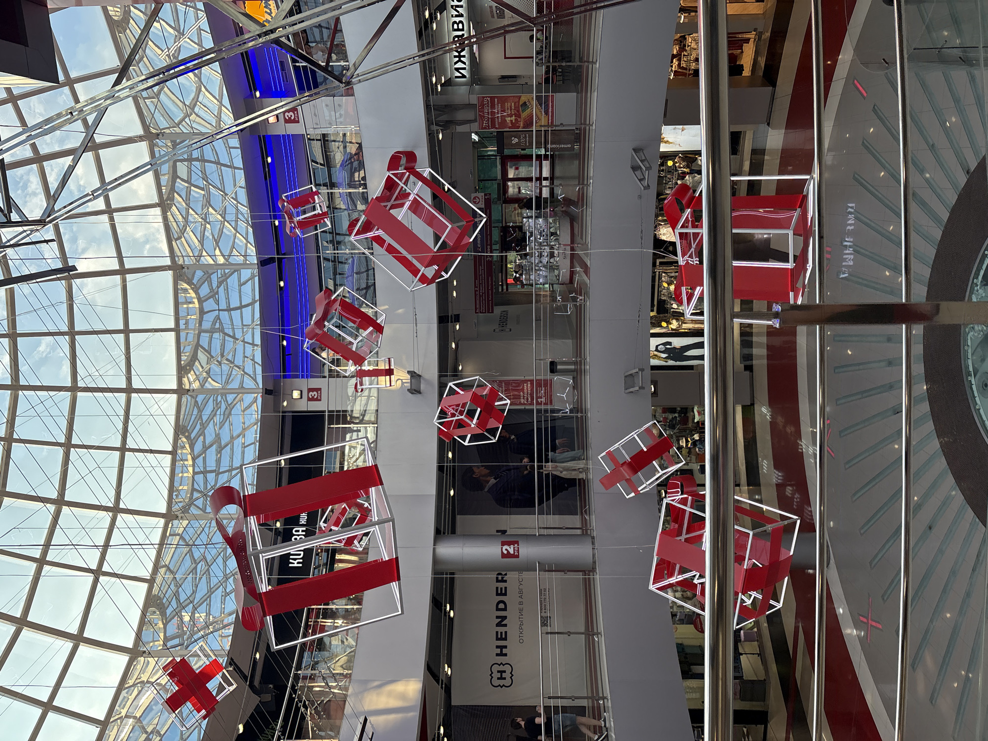 This image shows the interior of a multi-level shopping mall in Izhevsk, Russia. The mall features a large, domed glass ceiling and is decorated with large, red and white cube-shaped gift box sculptures hanging from the ceiling. The mall floors are visible, with shops and escalators. People are present on the different levels, walking and browsing. The lighting appears to be a mix of natural light from the ceiling and artificial lighting from within the shops. The overall scene suggests a commercial and recreational space.