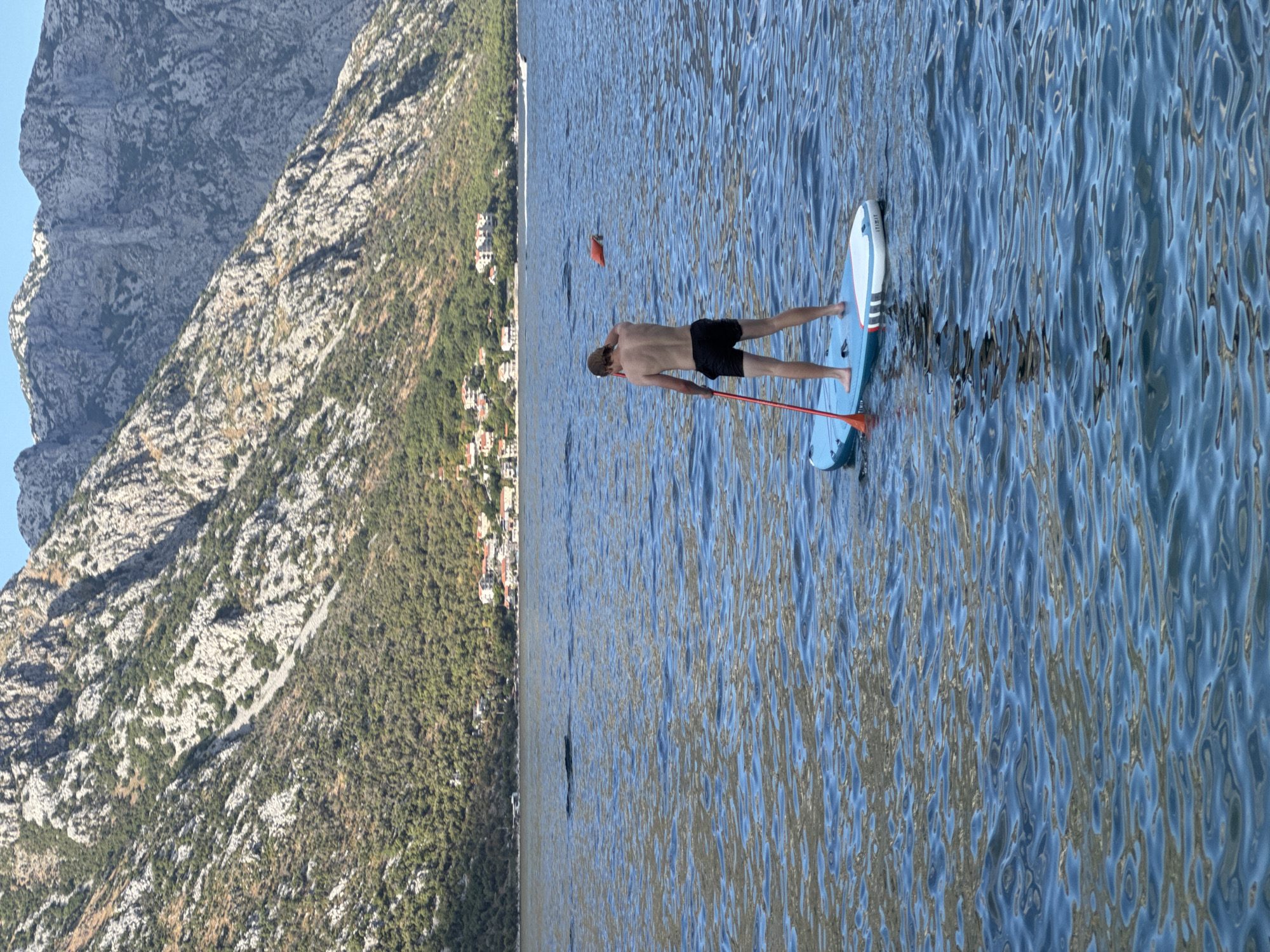In the city of Prčanj, Montenegro, a young person stands on a paddleboard in the calm, blue waters of a bay. The individual, wearing black shorts, is facing away from the camera, holding a red paddle. The paddleboard is light blue and white with red accents. In the background, a lush green hillside covered in trees rises steeply, dotted with red-roofed houses. Above the hill, jagged, rocky grey mountains dominate the skyline under a clear blue sky. The water is slightly rippled, reflecting the colors of the sky and the surrounding landscape. An orange buoy floats in the distance.