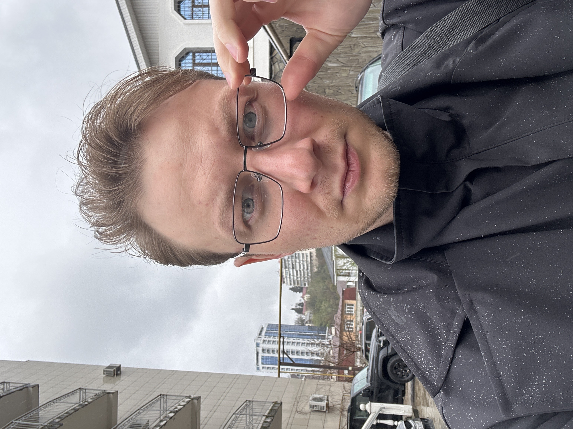 A medium shot depicts a fair-skinned, light-haired man holding eyeglasses in his right hand, facing the camera. He has a light beard and is wearing a black jacket. The background shows a cityscape, including apartment buildings and a taller high-rise building under an overcast sky. Based on architectural features and the high-rise building, the location is plausibly identified as being within Sochi, Russia. There appears to be light rain, with water droplets visible on the jacket. A black vehicle and a white object are also visible in the background street scene.