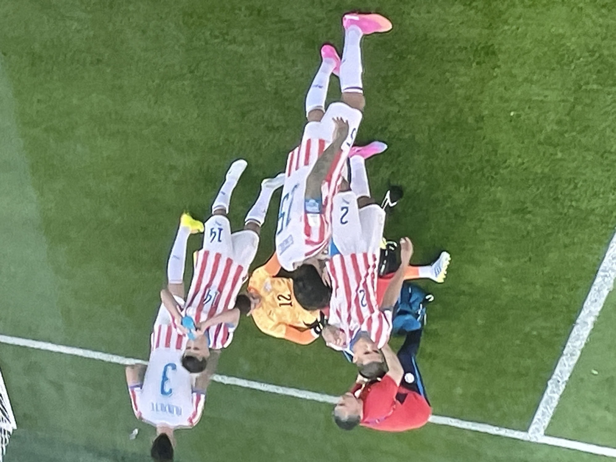 A group of male soccer players in red and white striped jerseys and white shorts are on a grassy field. One player, wearing a yellow jersey and black shorts, is lying on the ground. A man in a red shirt and black shorts, possibly a coach or trainer, is kneeling beside the player on the ground. Two other players in red and white striped jerseys are standing near the player on the ground, with one appearing to drink from a blue bottle. Another player in a red and white striped jersey with the number 3 on the back is standing further away. The scene appears to be a soccer match taking place on a well-maintained grass field, likely at a stadium given the markings on the field. The overall atmosphere suggests a pause in the game due to an injury. The image does not contain enough information to determine its location in the city of Zhukovo, Russia.