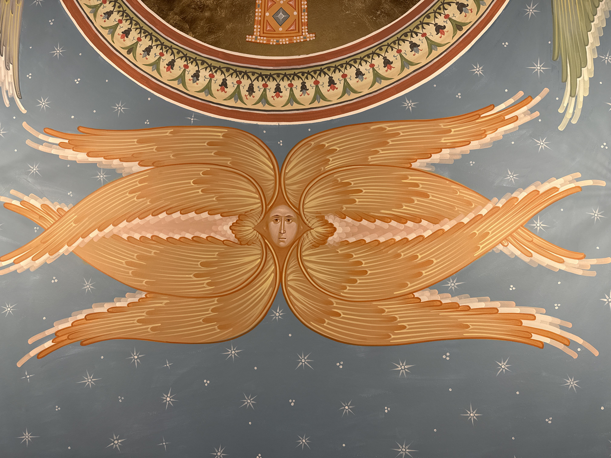 The image depicts a fragment of a religious mural, likely inside a church. The central focus is an angel with six wings emanating from a face. The angel and wings are painted in shades of orange and white with detailed line work suggesting feathers. The background is a blue-grey color dotted with stylized white stars of varying sizes and small clusters of white dots. Above the angel is a semi-circular band with a decorative border of floral and geometric motifs, with a gold-colored element containing a diamond-shaped icon above it. It is difficult to assess whether it is located in Saint Petersburg, Russia without external information.