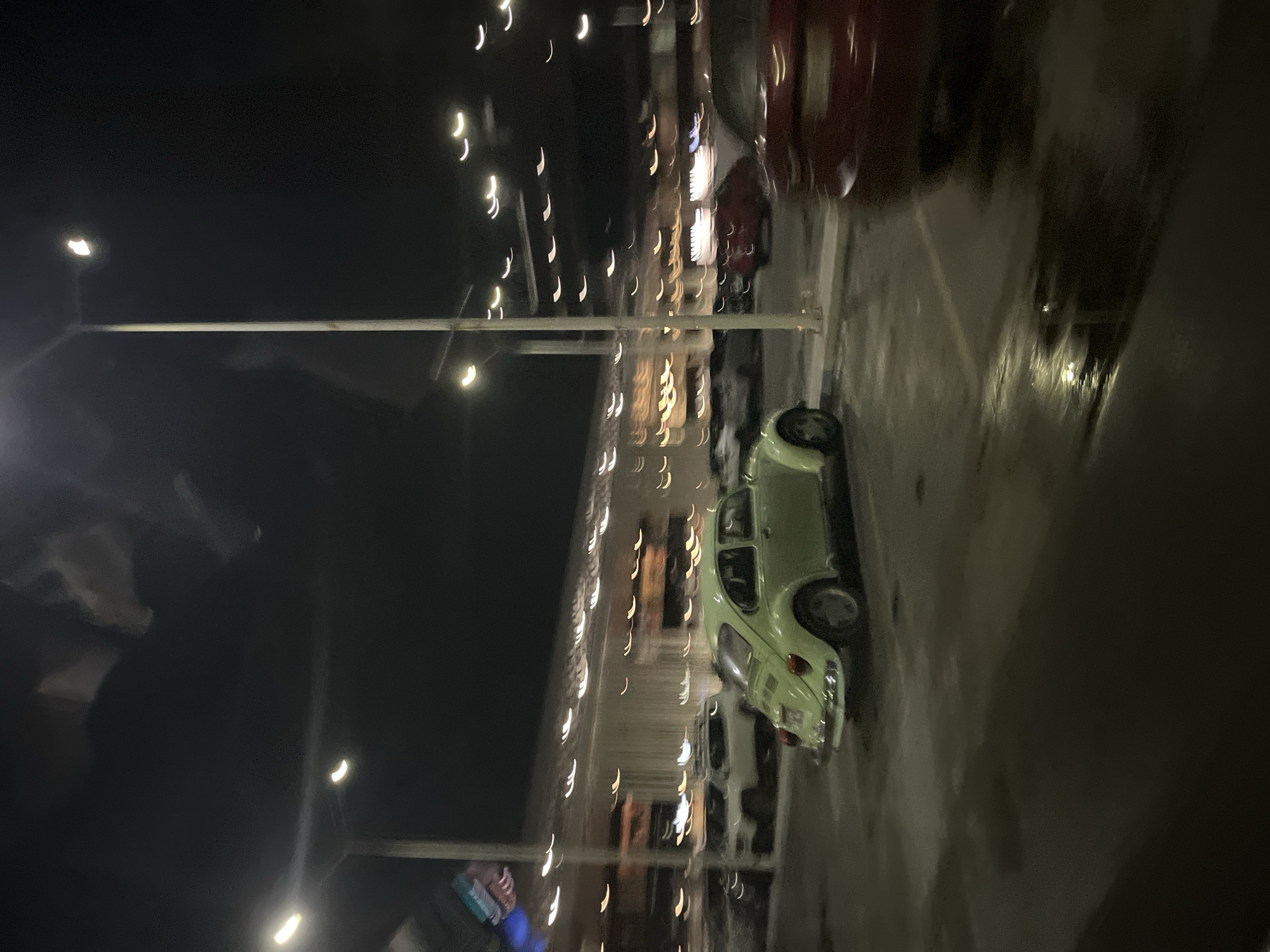 At night in Puebla, Mexico, a pale green Volkswagen Beetle is parked in a lot with wet pavement. Streetlights line the left side of the image, along with rows of blurred storefronts. A red car is parked behind the Beetle. Person, with blue and white jacket is in the left corner.