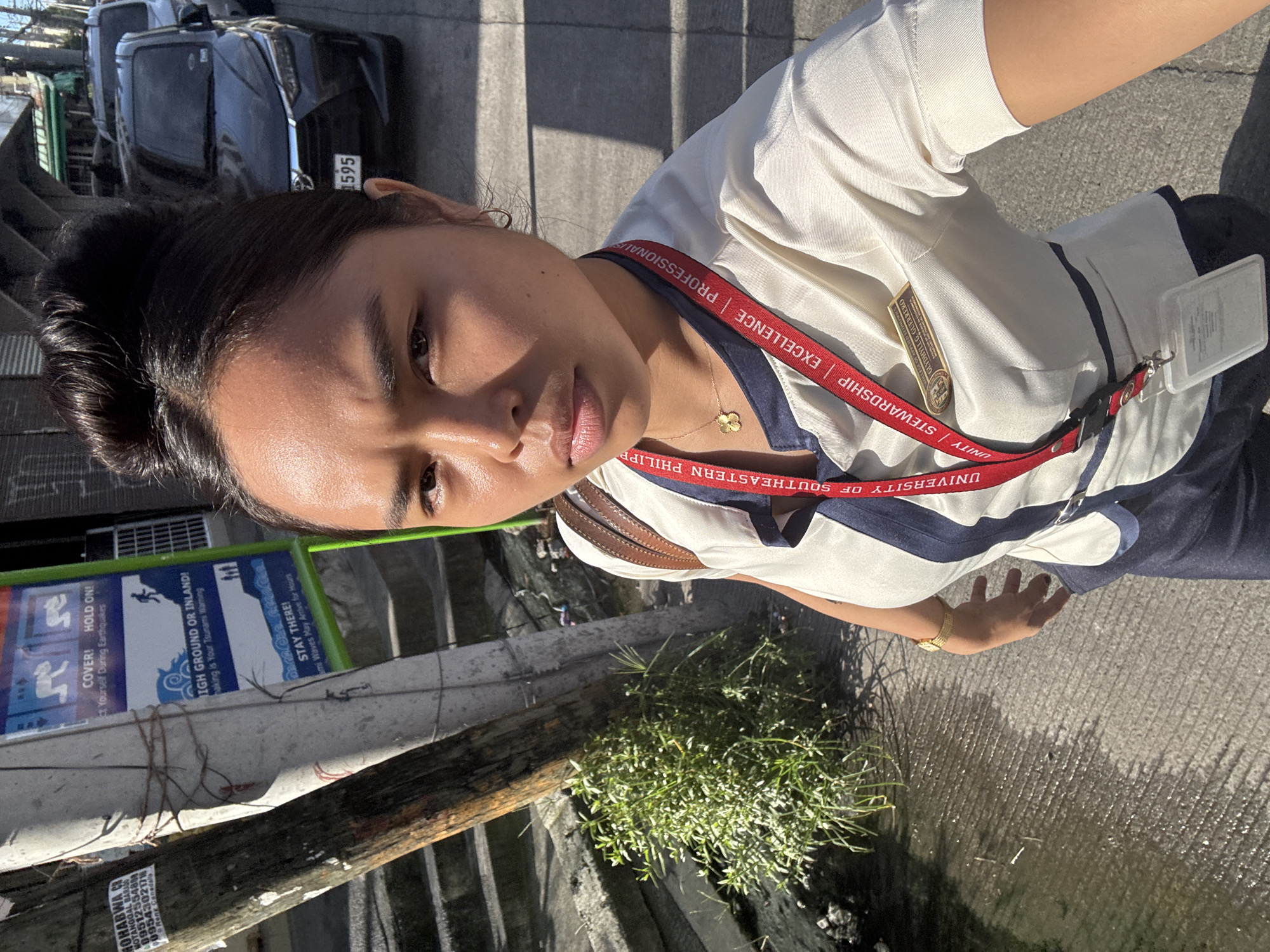 A young woman is taking a selfie outdoors in what appears to be an urban environment. She is wearing a light-colored uniform shirt with a dark blue collar and trim, adorned with a red lanyard and a school ID. The lanyard has text that appears to read "UNIVERSITY OF SOUTHEASTERN PHILIPPINES" and "STEWARDSHIP EXCELLENCE PROFESSIONALISM". A name tag on her shirt reads "GLYDIL G. GUERRERO". She has her hair styled in a bun and is wearing a gold necklace with a flower pendant and gold hoop earrings.
Behind her, a gray concrete pole is visible, wrapped with twine and bearing several stickers. To the left of the pole, a wooden pole is also visible, covered in more stickers. Further back, a set of concrete stairs leads upwards. A green and blue sign is mounted on the concrete pole, displaying safety instructions with text such as "COVER! HOLD ON!", "HIGH GROUND OR INLAND!", and "STAY THERE!".
To the right of the woman, a black car with a license plate ending in "1595" is parked on the side of the road. Another car is partially visible behind it. The ground surface is a textured concrete sidewalk. A small cluster of green plants grows near the base of the poles. The lighting suggests it is daytime, with strong sunlight casting shadows. The overall scene depicts a person in a uniform, possibly a student or employee, standing on a sidewalk in what is identified as Davao City, Philippines.