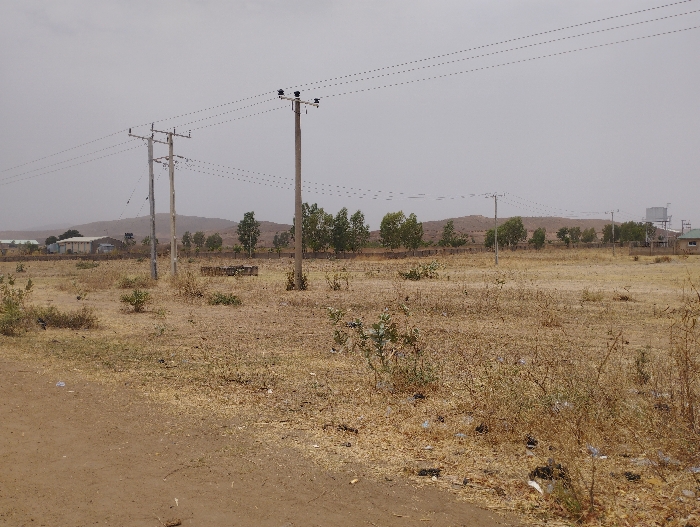 The image shows a dirt road in a rural area, with a water tower visible in the distance. Two electricity poles stand on either side of the road, with a network of power lines strung between them. The ground is dry and sparsely vegetated, with scattered shrubs and small trees. The background is dominated by a hill range, which is visible behind the power lines. The sky is overcast, and the overall impression is of a desolate and somewhat barren landscape. The scene is in the city of Kwadon, Nigeria.