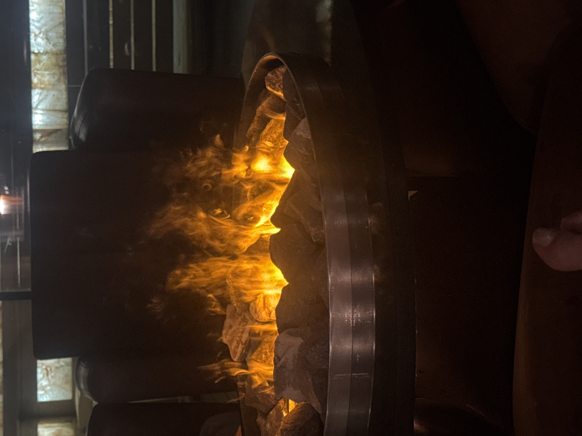 The image shows a close-up of a modern, round fire pit filled with rocks and flames. The fire pit is situated in a dimly lit, indoor setting, with dark, upholstered furniture visible in the background. The flames are bright orange and yellow, illuminating the rocks and casting a warm glow. The scene is captured from a low angle, focusing on the fire and the surrounding rocks. The overall impression is one of warmth and coziness. No specific location in Plovdiv, Bulgaria, can be identified from the image alone.